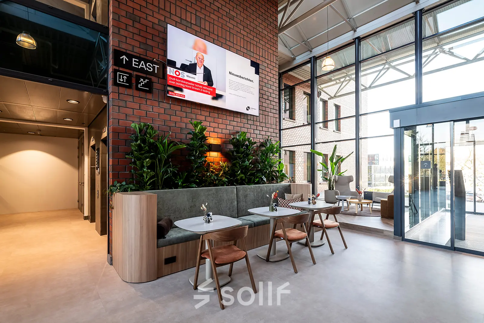 Modern office space at Hambakenwetering 5 in Den Bosch with stylish furnishings, indoor plants, and a wall-mounted screen. Perfect for office space rental, it features natural lighting and a comfortable seating area.