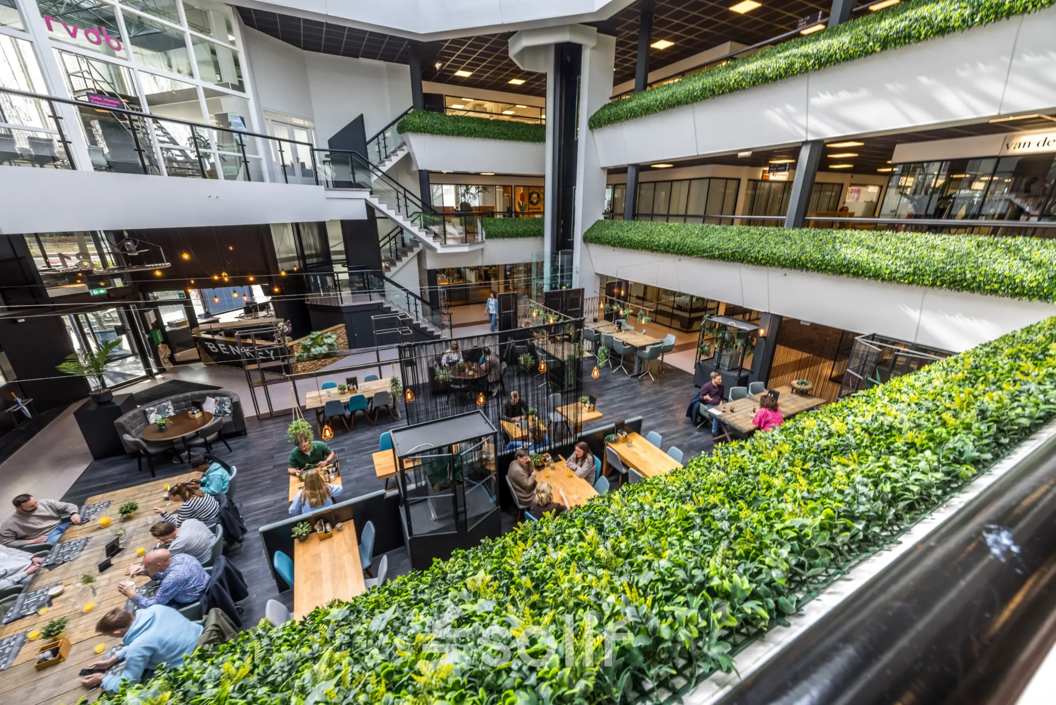Open-plan workspace at Eindhoven Airport's Luchthavenweg 81, featuring people collaborating at shared tables surrounded by greenery.