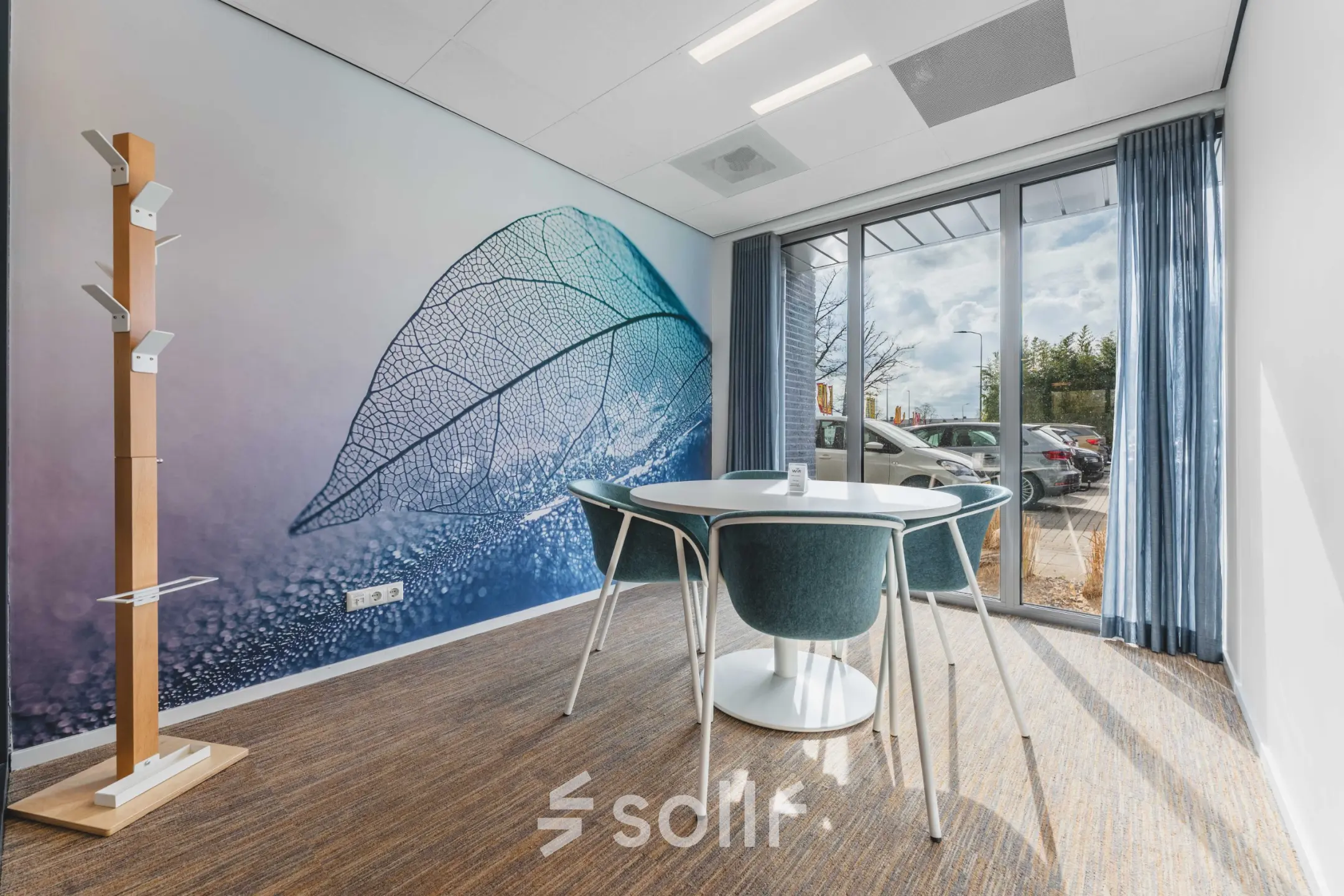 Bright office space with a large window, blue curtains, a wall mural, round table, and chairs for office space rental.