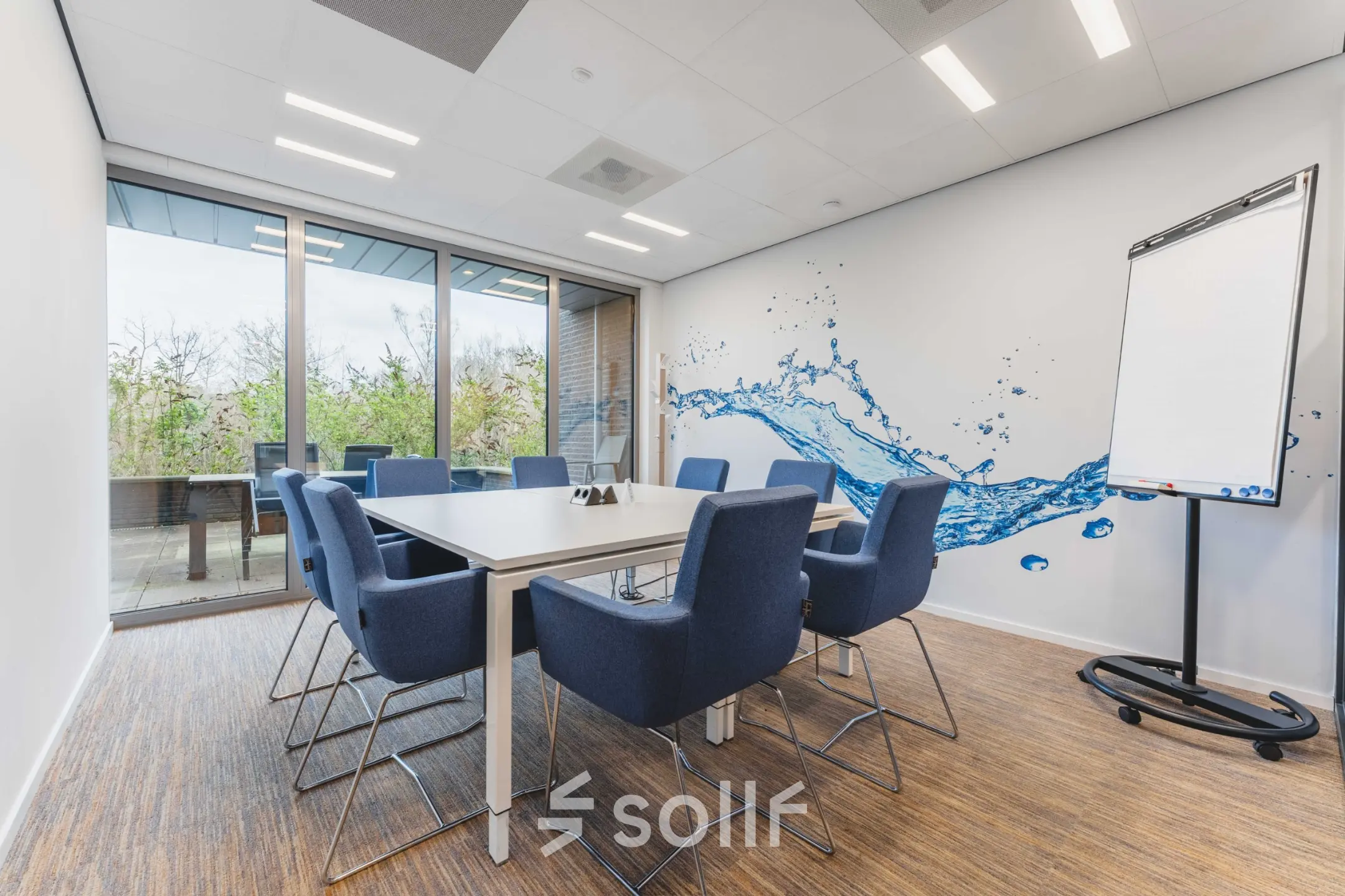 A modern conference room with a large table, blue chairs, and a glass wall in an office space available for rent at Hurksestraat 60, Eindhoven De Hurk.
