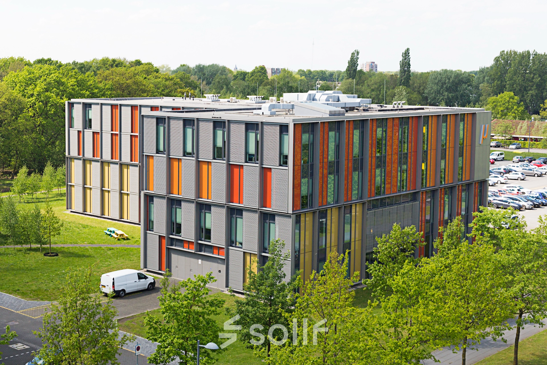 Exterior view of a modern, colorful office building at High Tech Campus 10, Eindhoven, surrounded by greenery with parked vehicles nearby. An ideal location for office space rental.