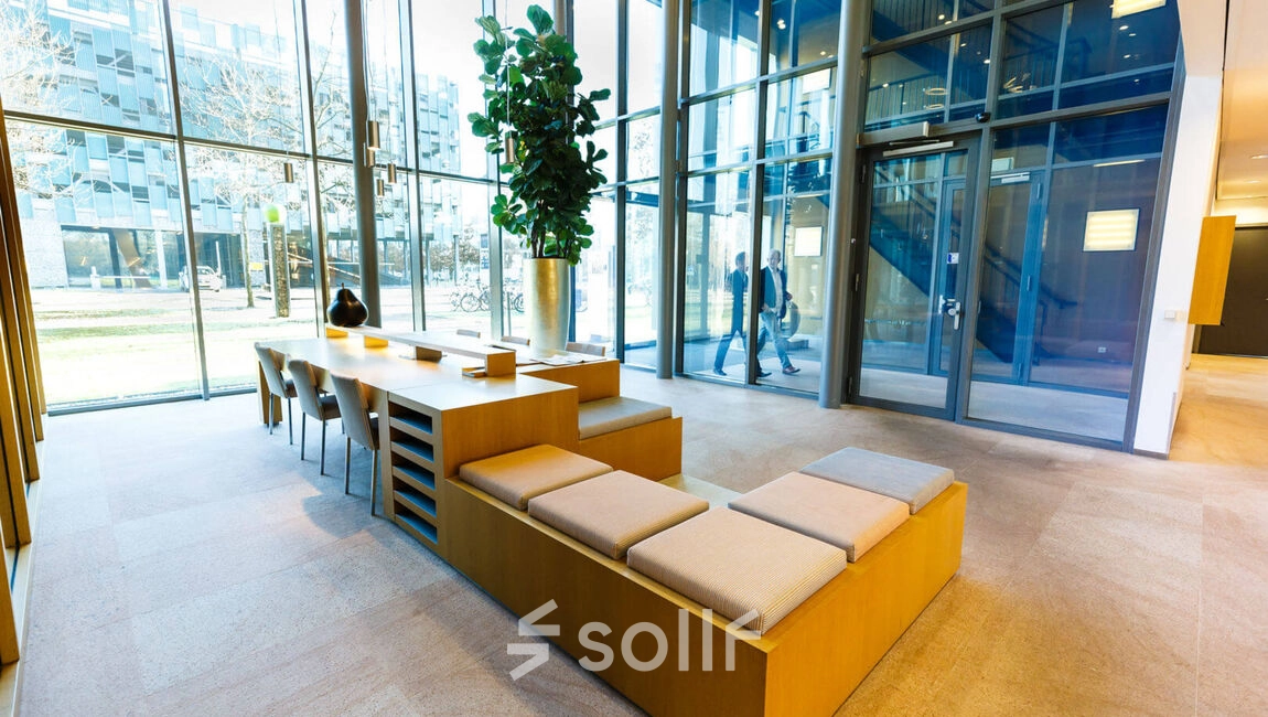 Modern office lobby at High Tech Campus 9, Eindhoven, with large windows, sleek wooden seating, and lush greenery. Ideal for businesses seeking office space rental.