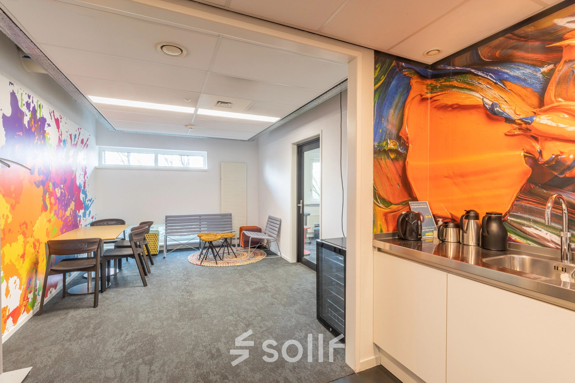 Vibrant office lounge at TU campus, De Zaale 11, Eindhoven. Features colorful mural walls, seating area with chairs and tables, and kitchenette. Ideal for office space rental enthusiasts seeking a dynamic environment.