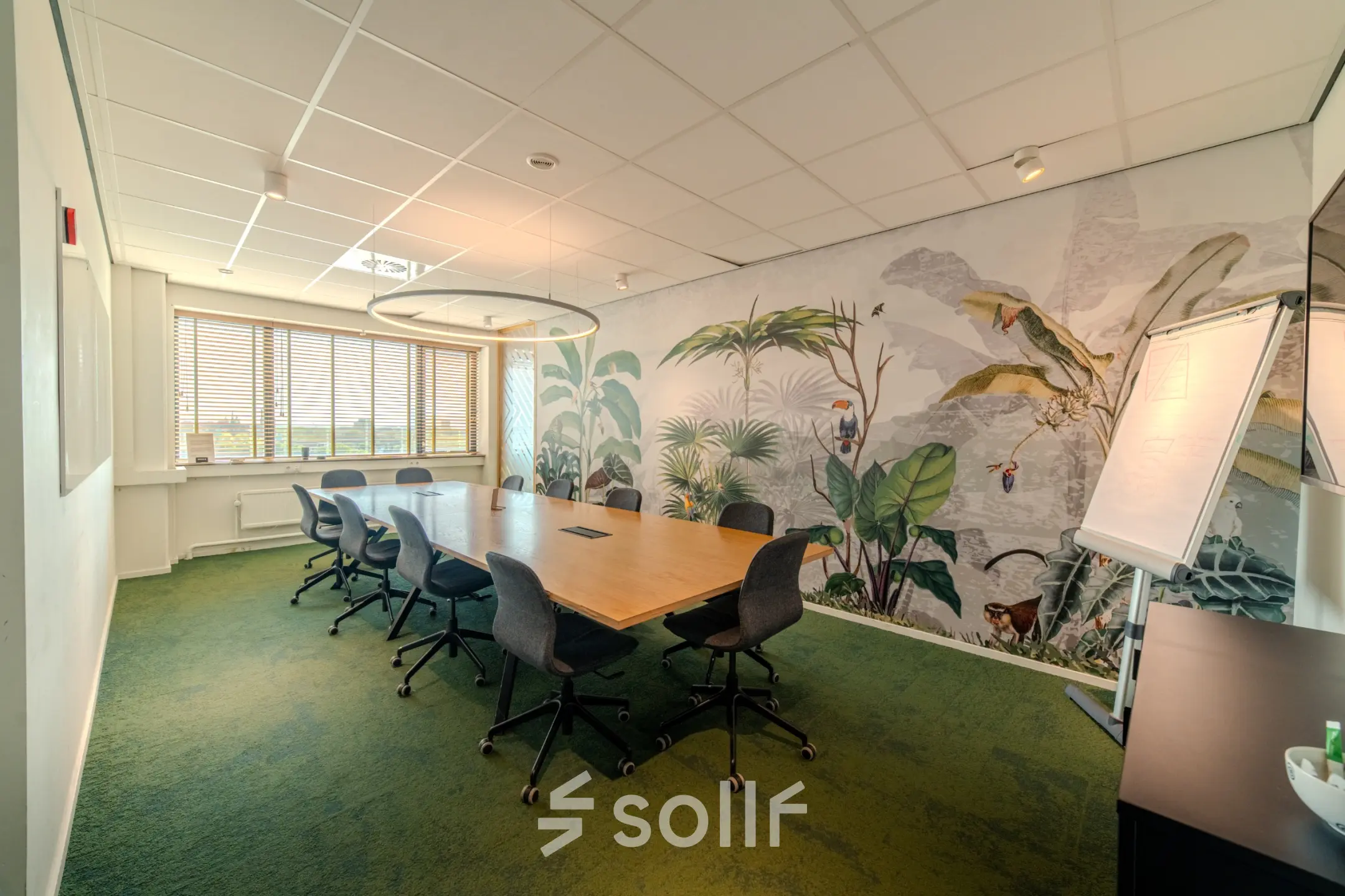 Bright meeting room with a large wooden table, green carpet, and tropical-themed wall mural, ideal for office space rental in Eindhoven Strijp-S.