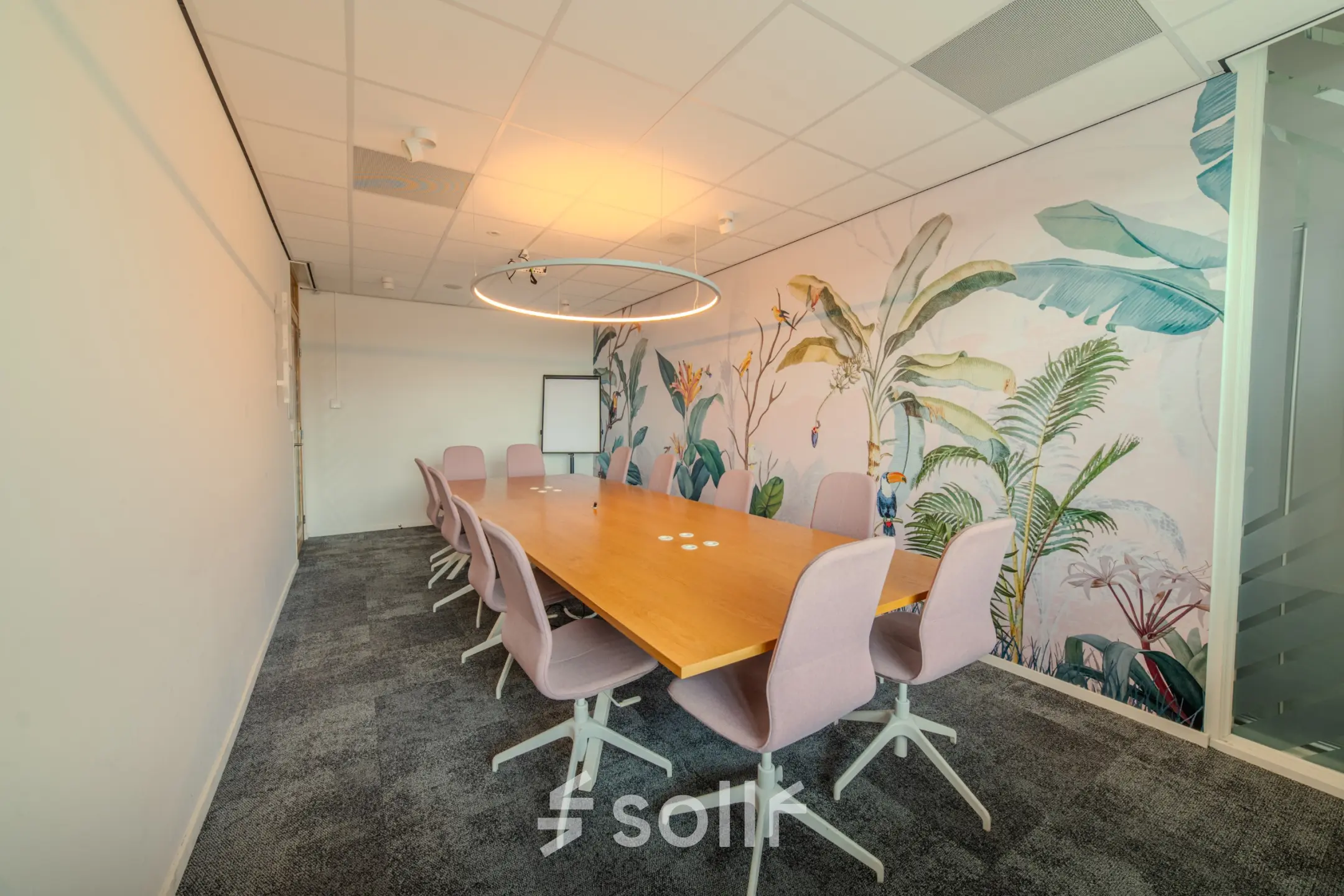 Stylish office space for rent with a long wooden table and modern chairs, featuring a wall with vibrant botanical art. Ideal for meetings or collaborative work.