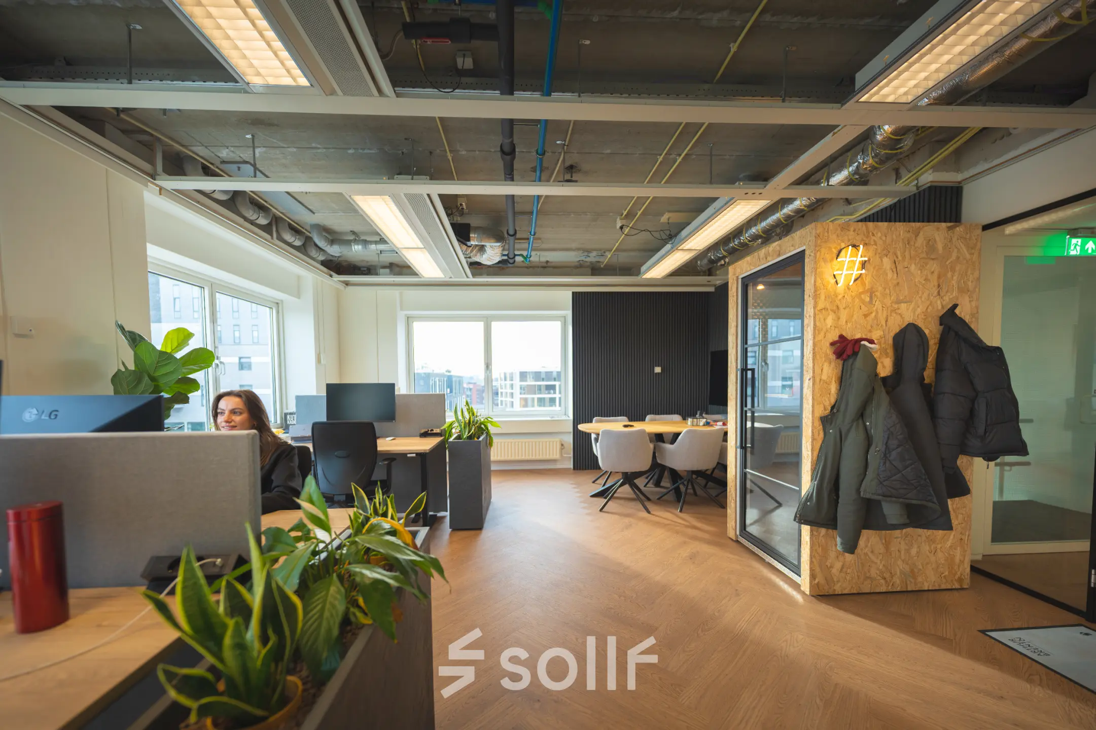 Modern office space at Brouwerijstraat 1, Enschede with contemporary furnishings, plants, and a glass-walled meeting area. A person is working at a desk, showcasing available office space rental options.