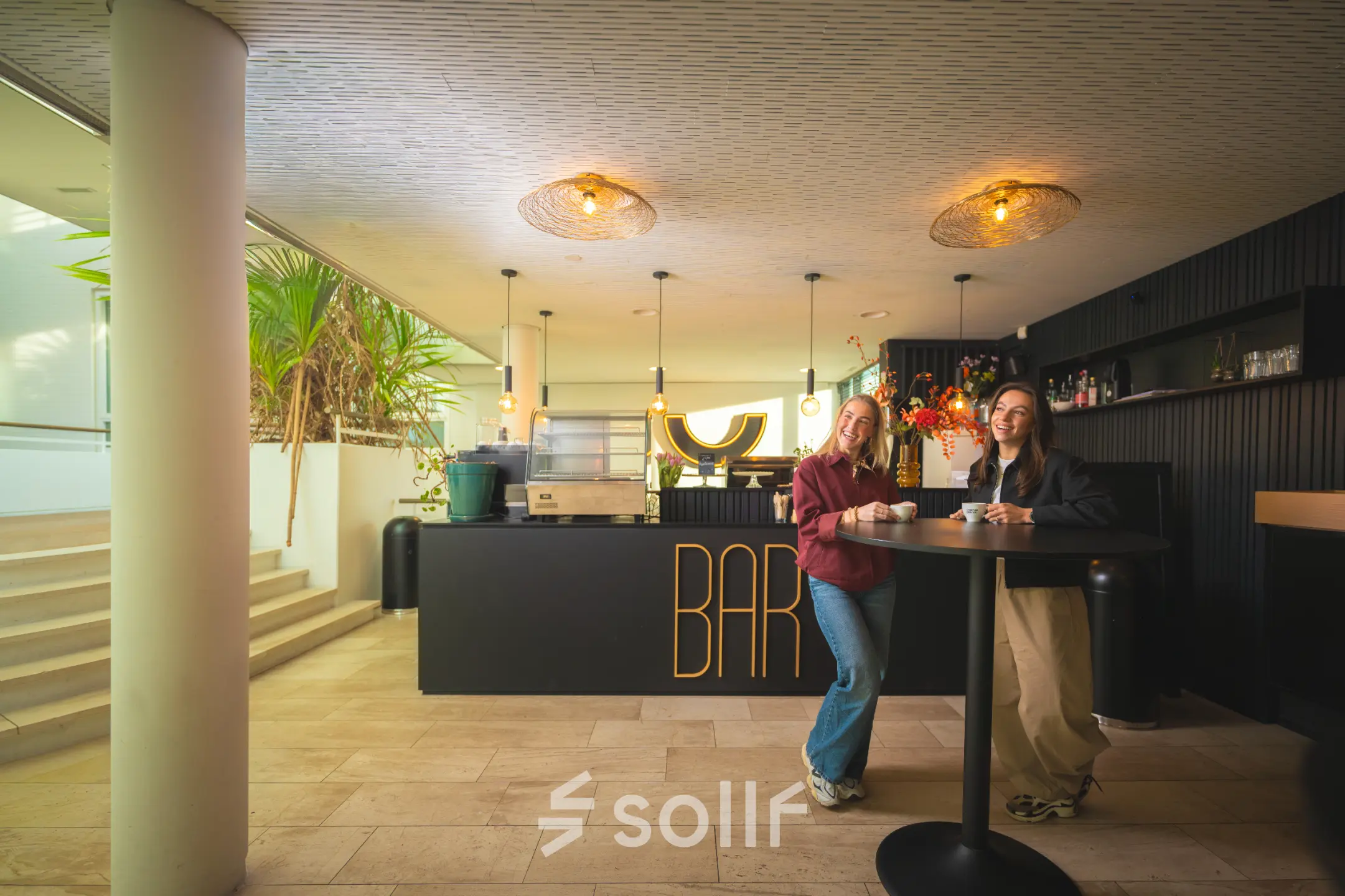Two people enjoying drinks at a modern bar area near stairs, located in Brouwerijstraat 1, Enschede, ideal for office space rental.