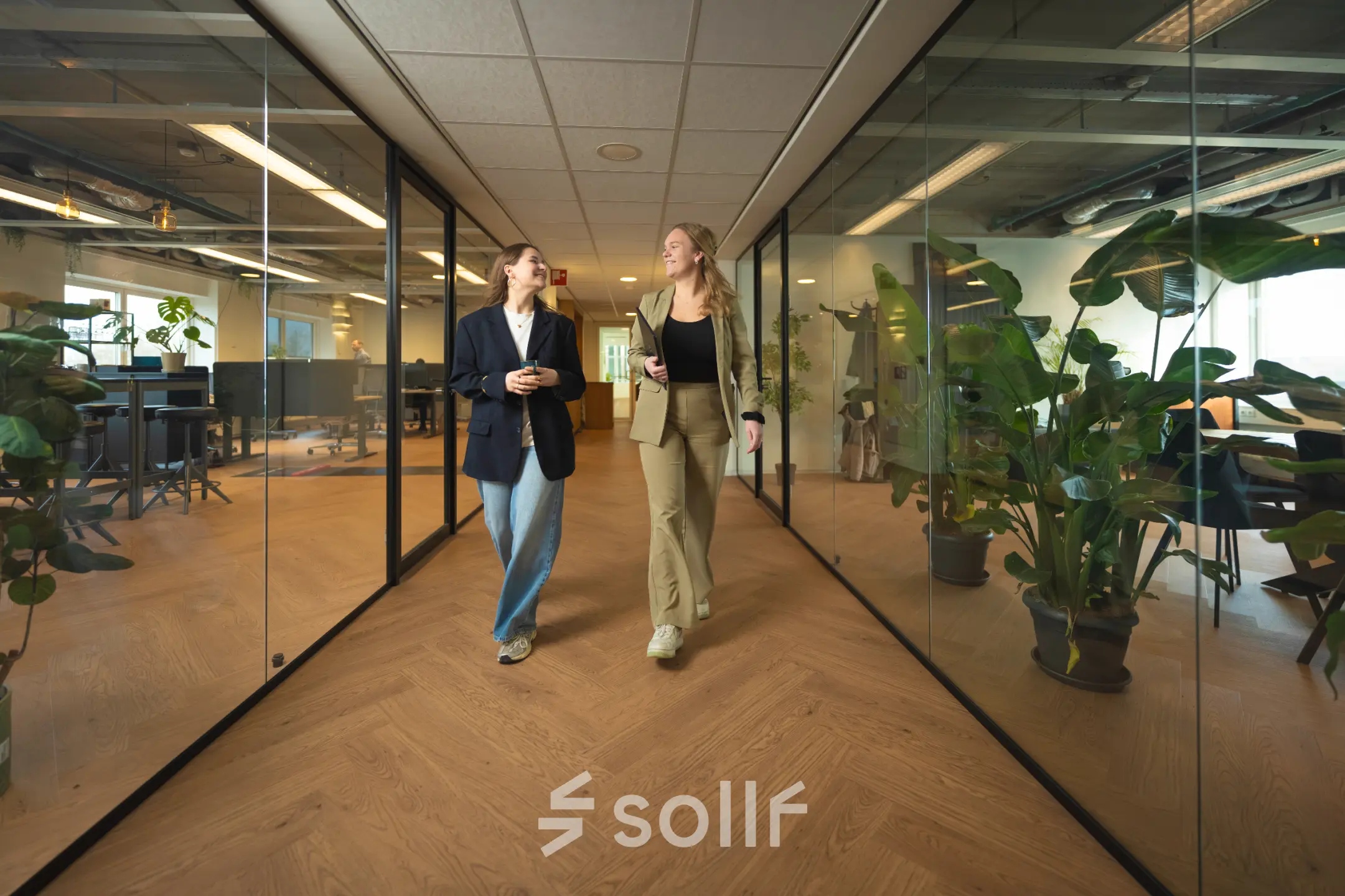 Two people walking and conversing in a modern office to rent at Brouwerijstraat 1, featuring glass walls and wooden flooring with office plants.