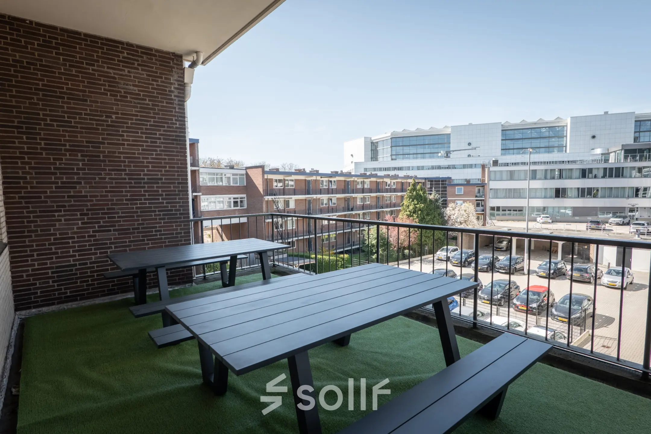 Outdoor office space rental at Kuipersdijk 2-4 in Enschede, featuring two black picnic tables on artificial grass, overlooking a parking area and adjacent buildings.