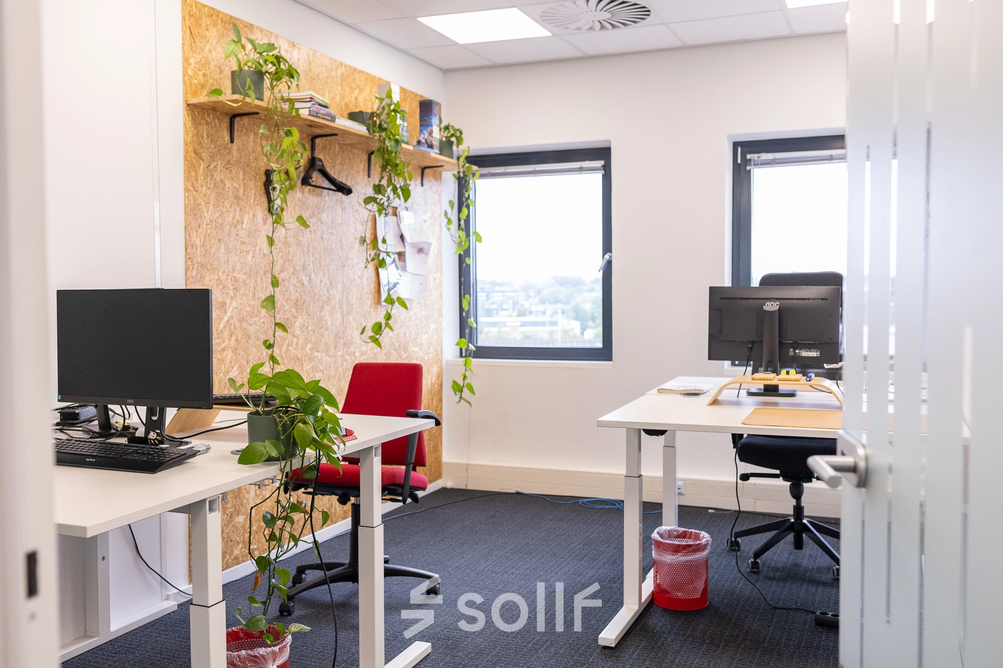 Bright, furnished office space in Gouda with desks and plants, ideal for office space rental. Neutral decor with large windows providing natural light and a view. Two red chairs add a pop of color.
