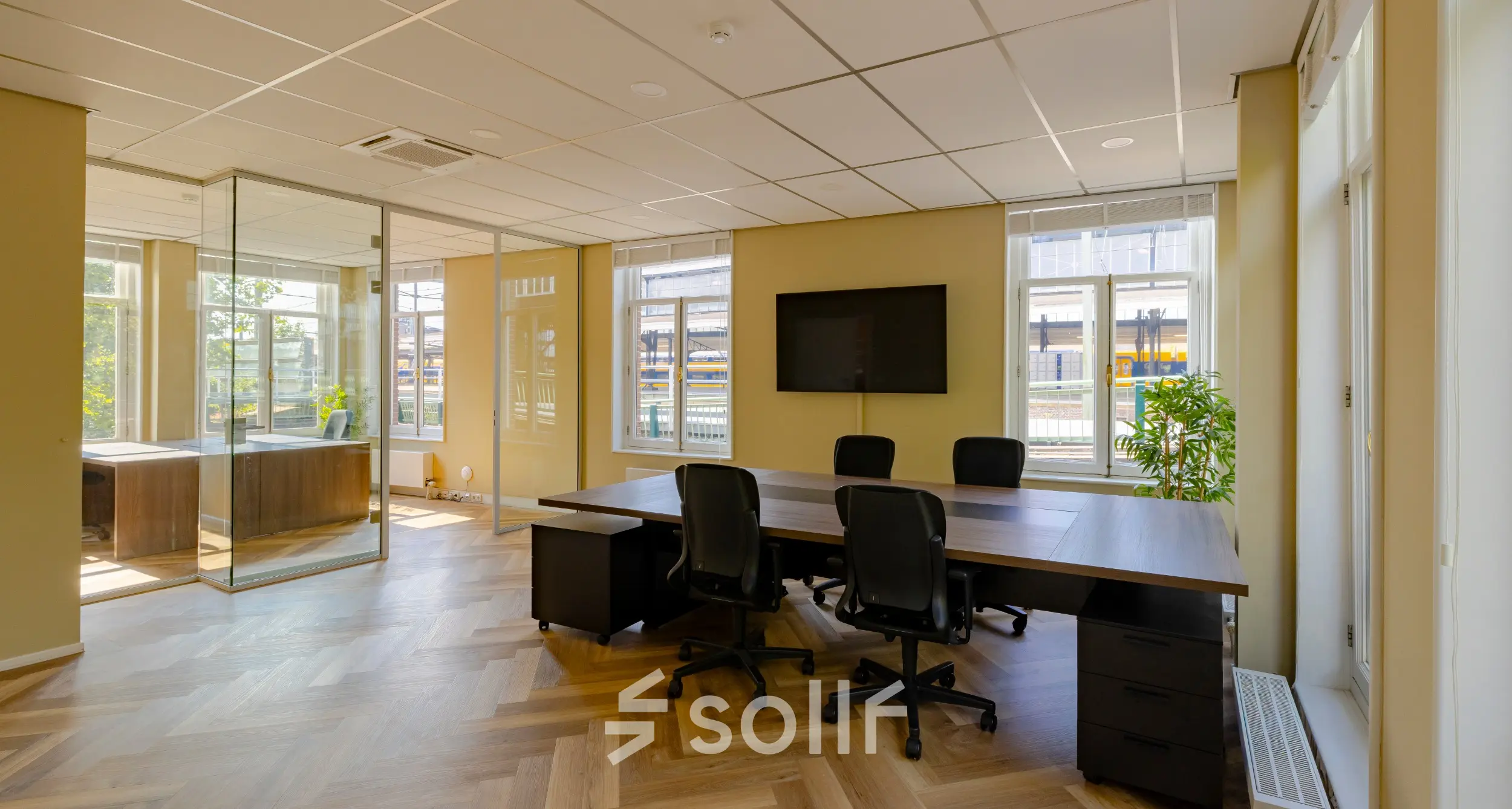 Bright office space rental at Kennemerplein 20, featuring a large wooden desk with chairs, glass partition, and modern decor; natural light fills the room through multiple windows.