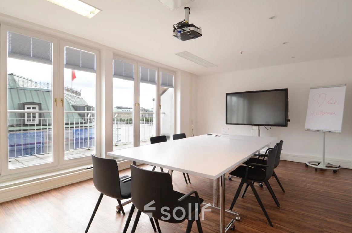 Bright office space with large windows, a table, chairs, and a TV, perfect for meetings. Ideal office space rental at Colonnaden 5, Hamburg Neustadt.