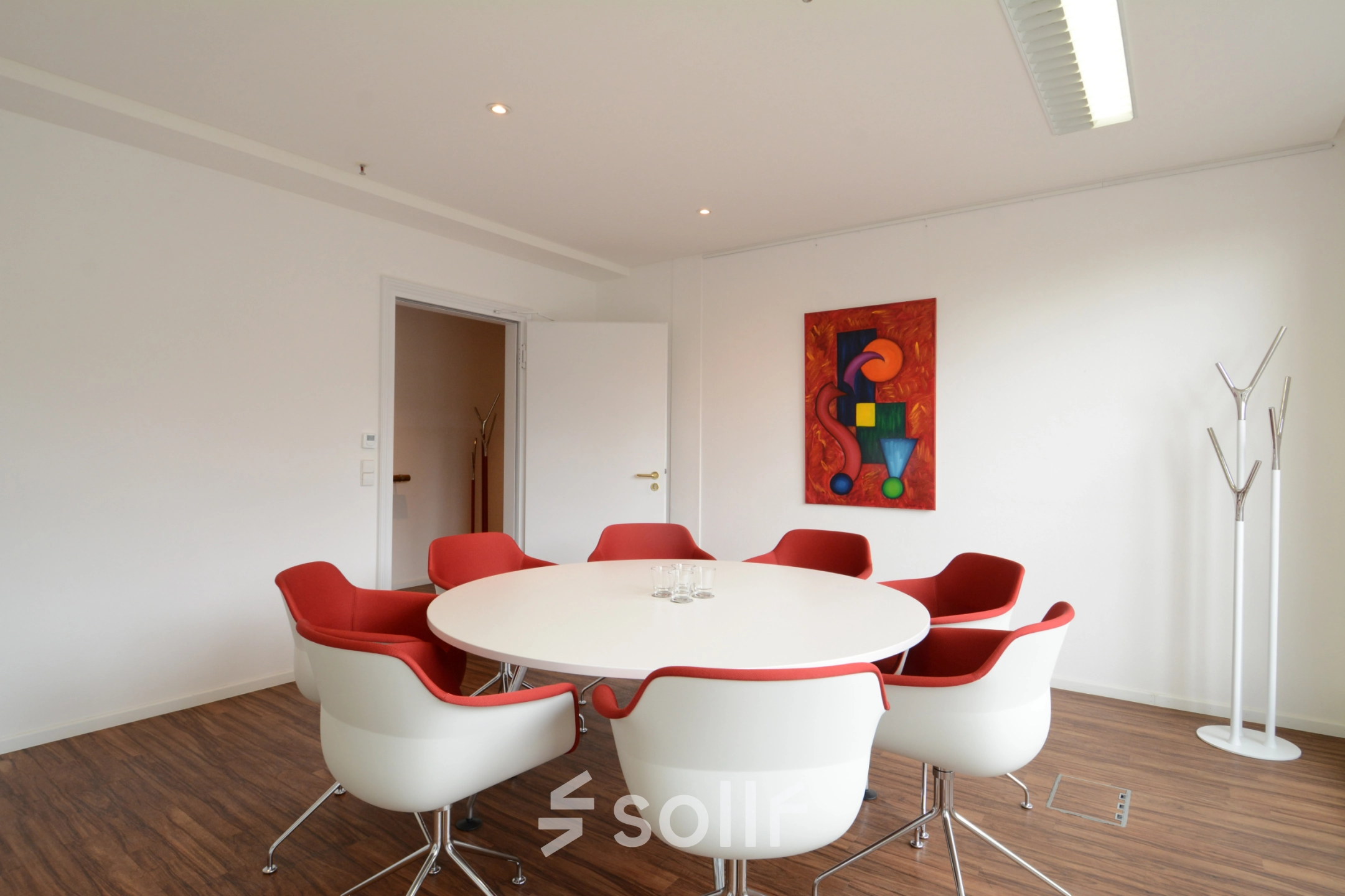 Bright office space with red chairs around a white table, ideal for meetings. Modern decor includes abstract art on the wall. Perfect for rent office space inquiries.