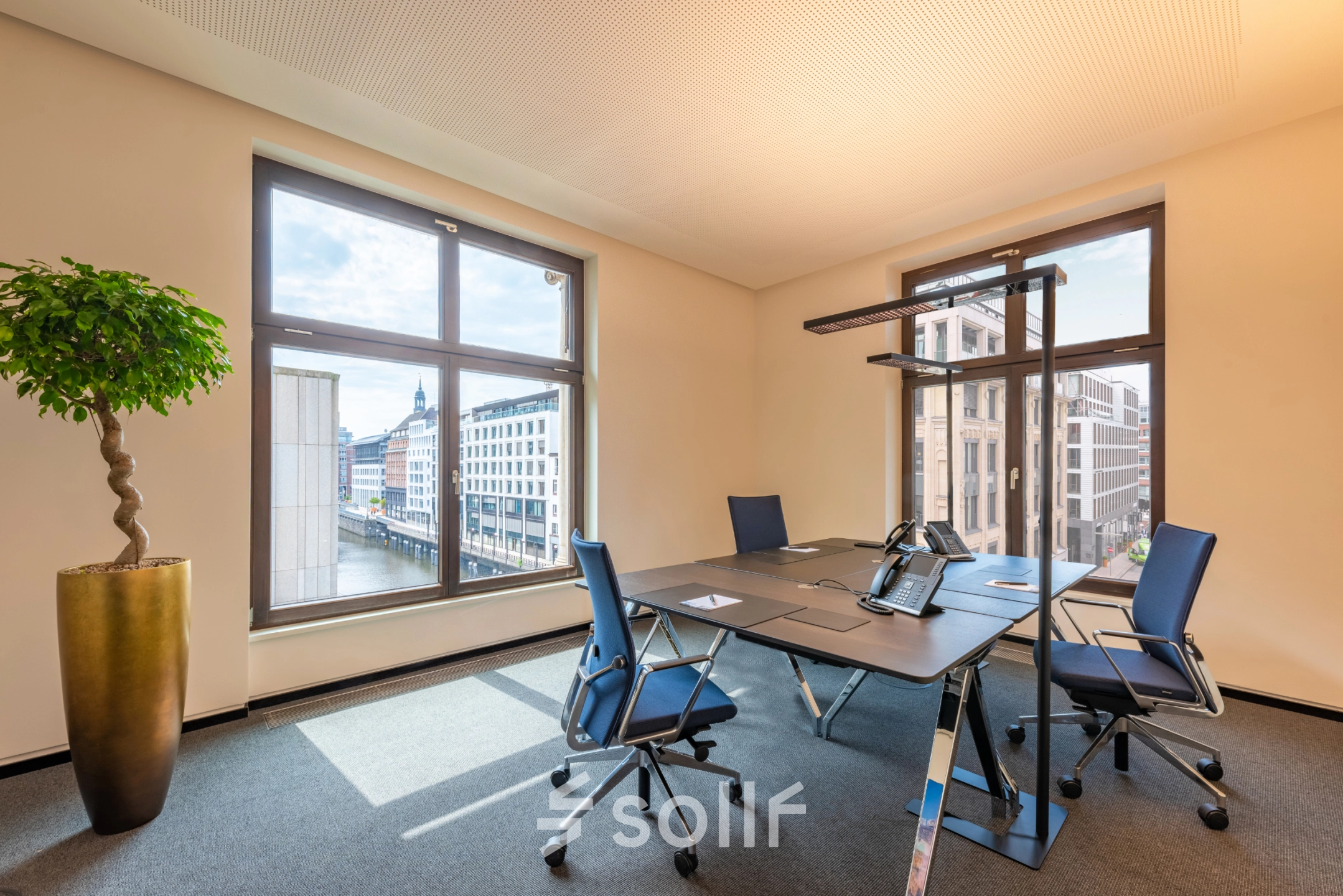 A bright, furnished office to rent at Alter Wall 32, Hamburg Old Town, featuring large windows, a meeting table, and chairs overlooking the cityscape.