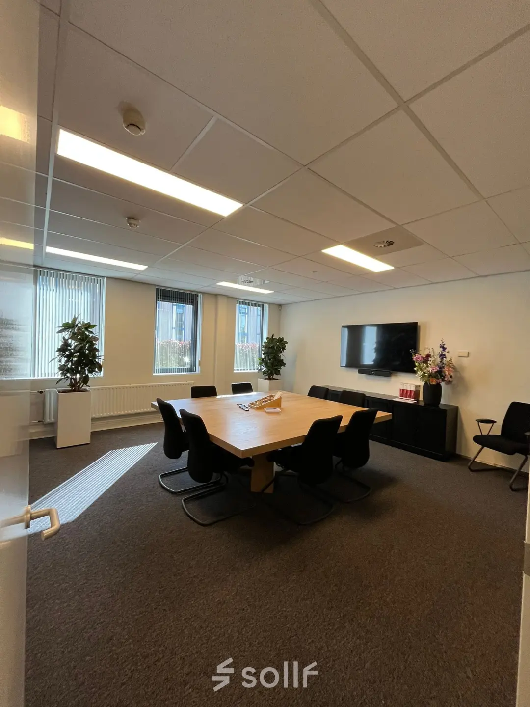 Modern office space in Hilversum with a large wooden table, surrounded by black chairs, suitable for meetings. Bright lighting and indoor plants add a fresh touch. Ideal for office space rental.