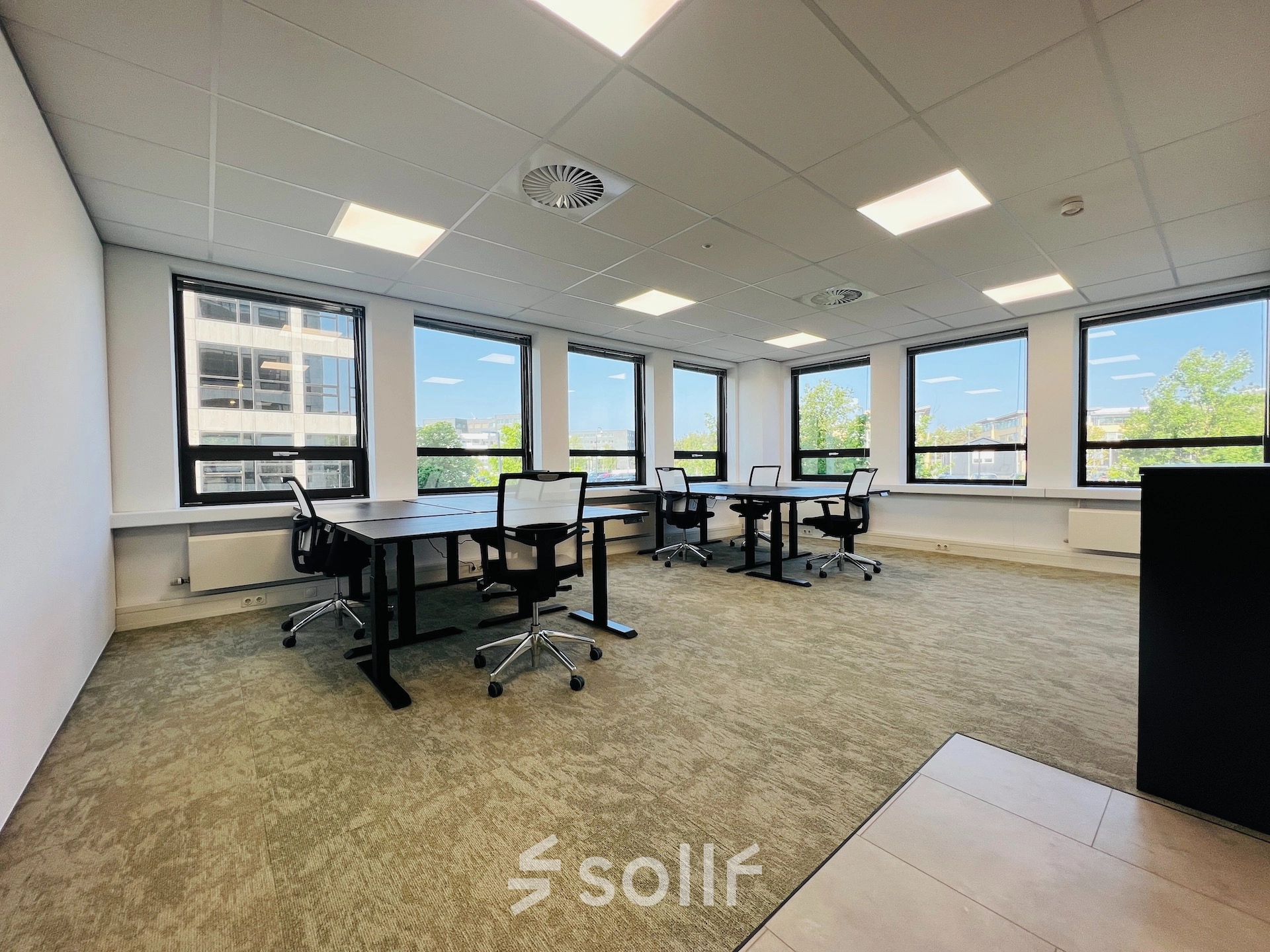 Bright office space in Hilversum featuring multiple black desks and mesh chairs by large windows with a view, available for rent.