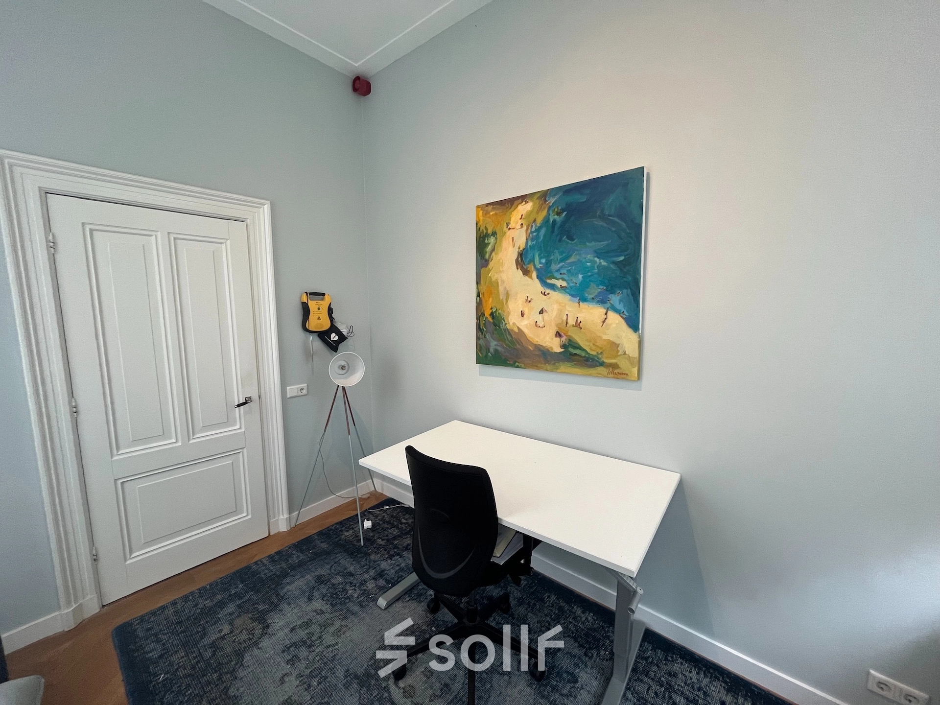 Small, furnished office space with a white desk, black chair, and colorful artwork on the wall, available for office space rental in Hilversum.