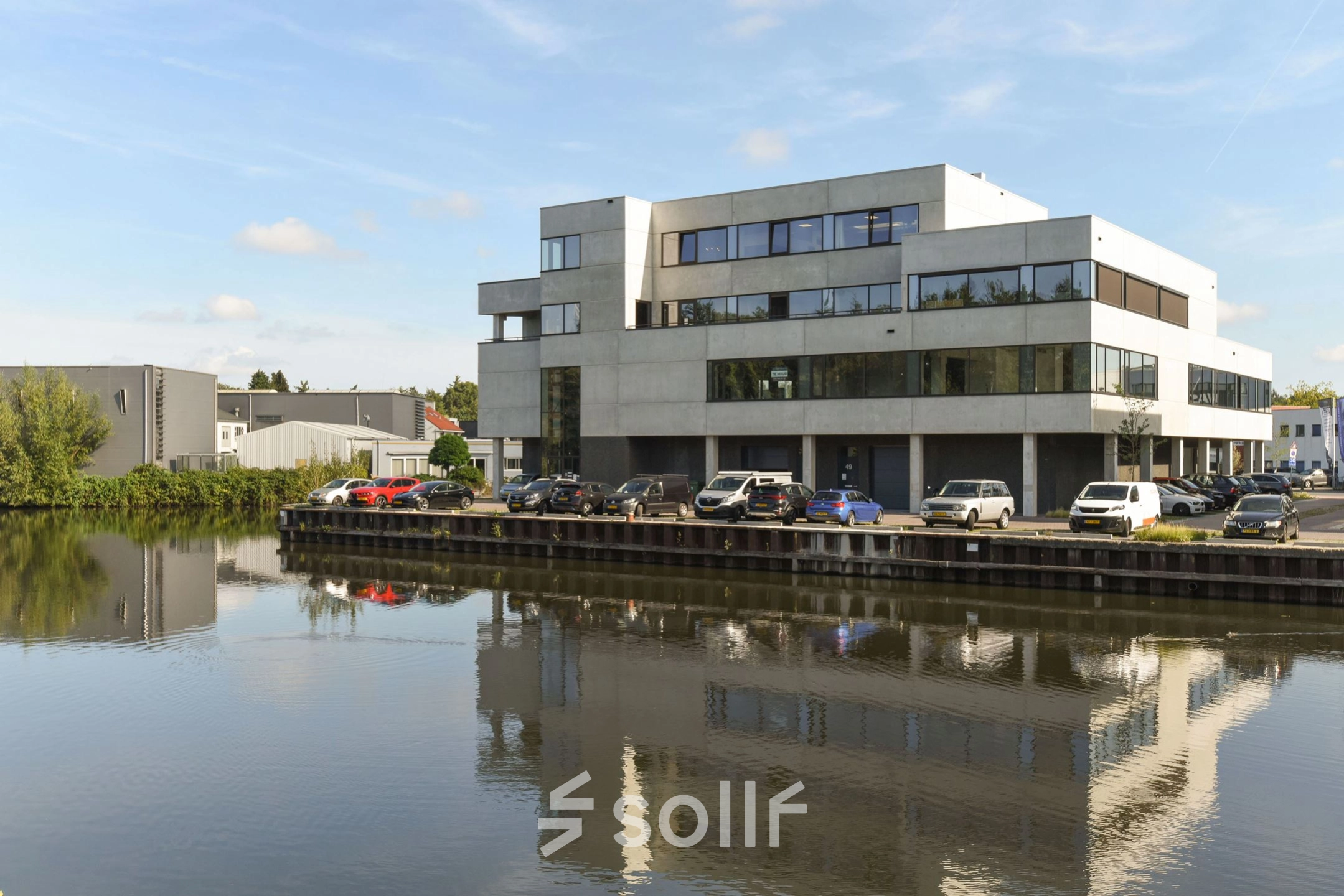 Exterior view of an office building at Zuiderloswal 55 in Hilversum, reflecting in a nearby canal. Multiple cars are parked outside. Rent office space in a scenic location.