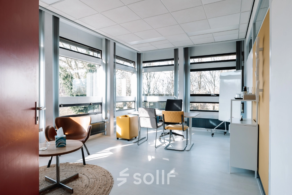 Bright office space with modern furnishings available for rent at De Vos van Steenwijklaan 75, Hoogeveen. Features large windows, stylish chairs, and a meeting table, ideal for office space rental.