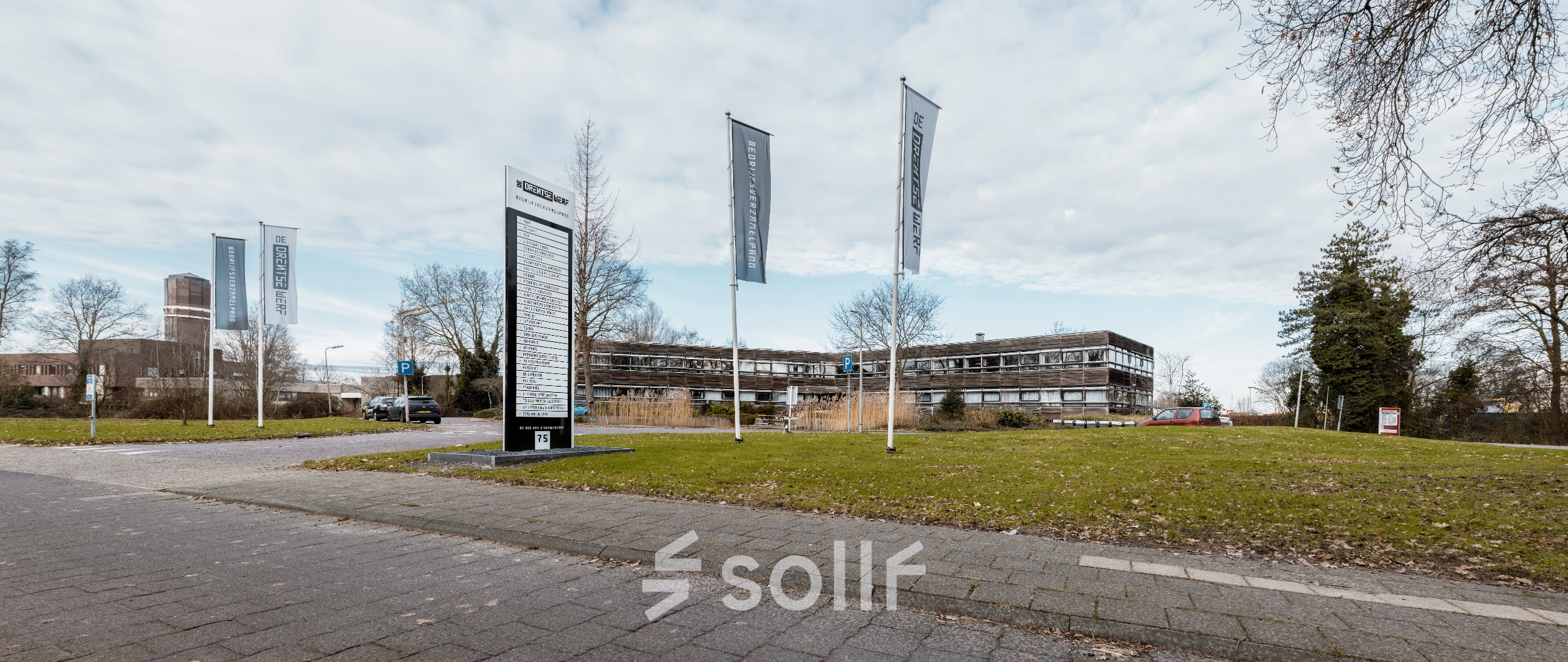 Exterior view of an office building at De Vos van Steenwijklaan 75, Hoogeveen, surrounded by greenery with signage and flags, offering office space rental options.