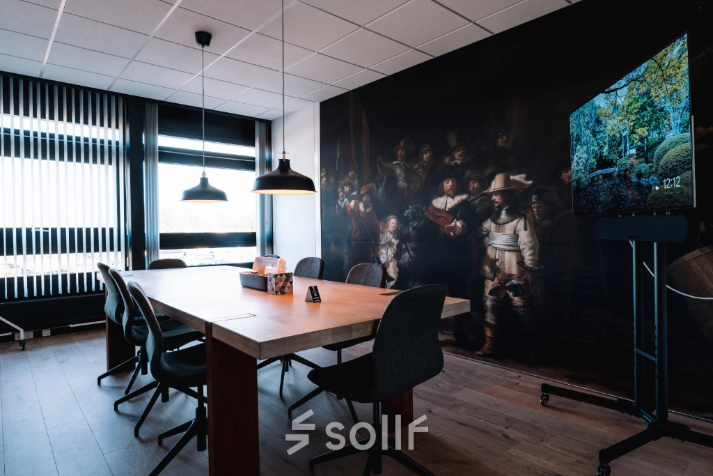 Cozy office space for rent in Hoogeveen featuring a large wooden table and modern chairs, ideal for meetings. A bright mural and a TV add a stylish touch to the room.