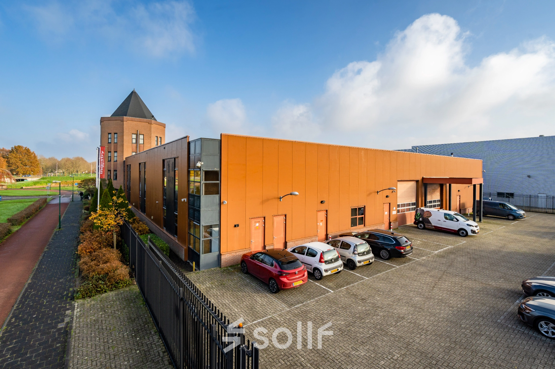 Exterior view of a modern office building at Hoofdveste 2, Houten, featuring an orange facade with a parking area in the foreground. Ideal for office space rental, this location offers convenience and accessibility.