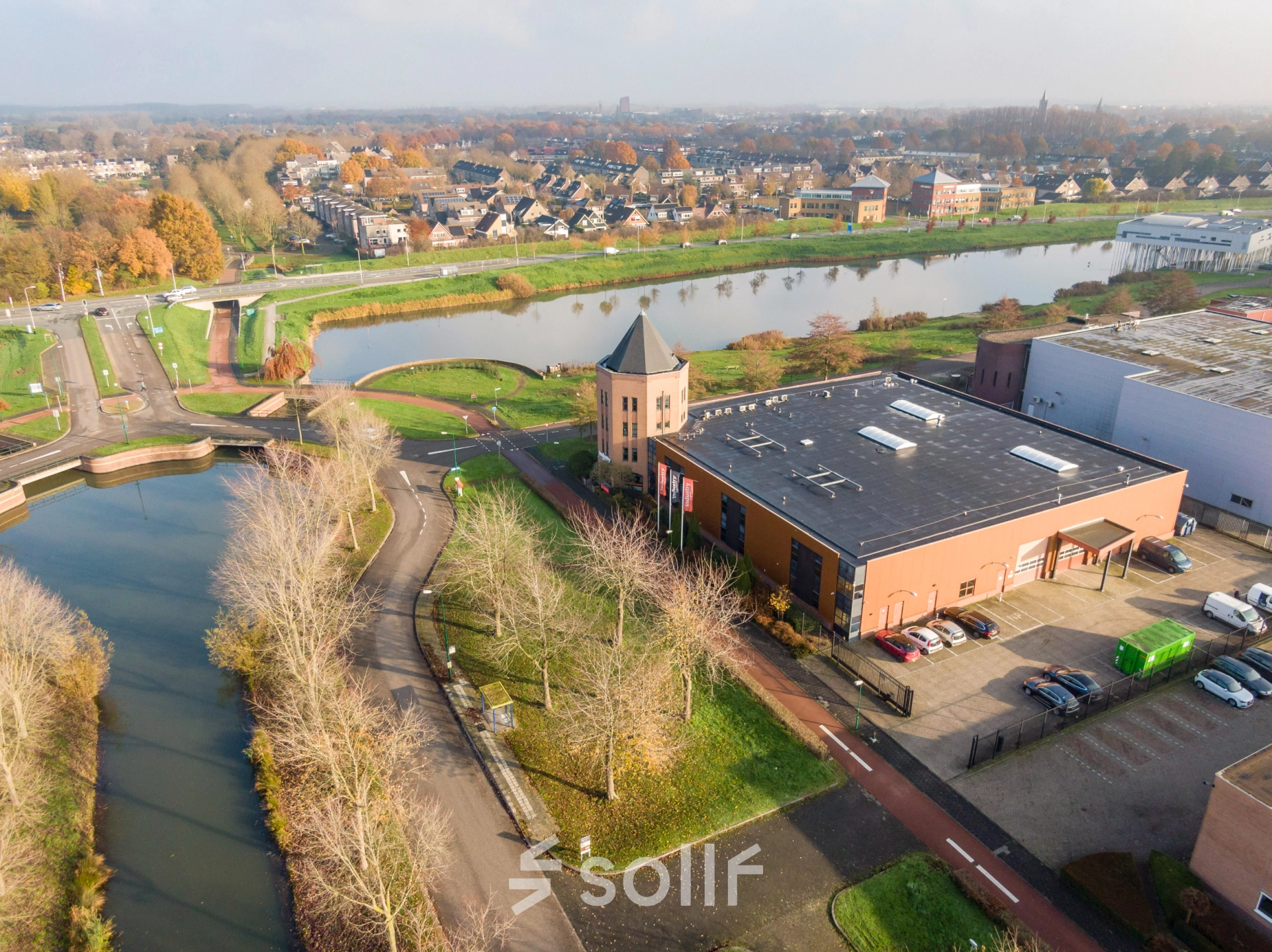 Aerial view of the office building at Hoofdveste 2, Houten, surrounded by a small canal and trees. The modern building offers office space rental in a scenic area with ample parking.