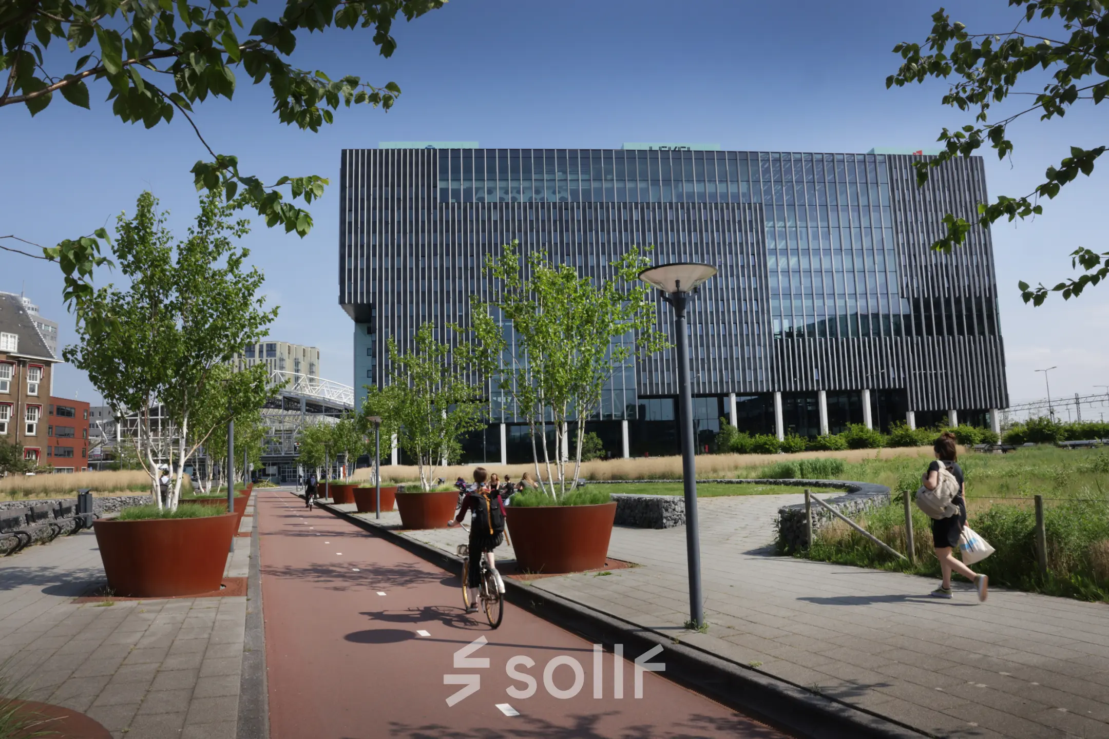 Exterior view of offices for rent at Bargelaan 200, Leiden, showcasing modern architecture with plant-lined walkways. People are walking and cycling outside.