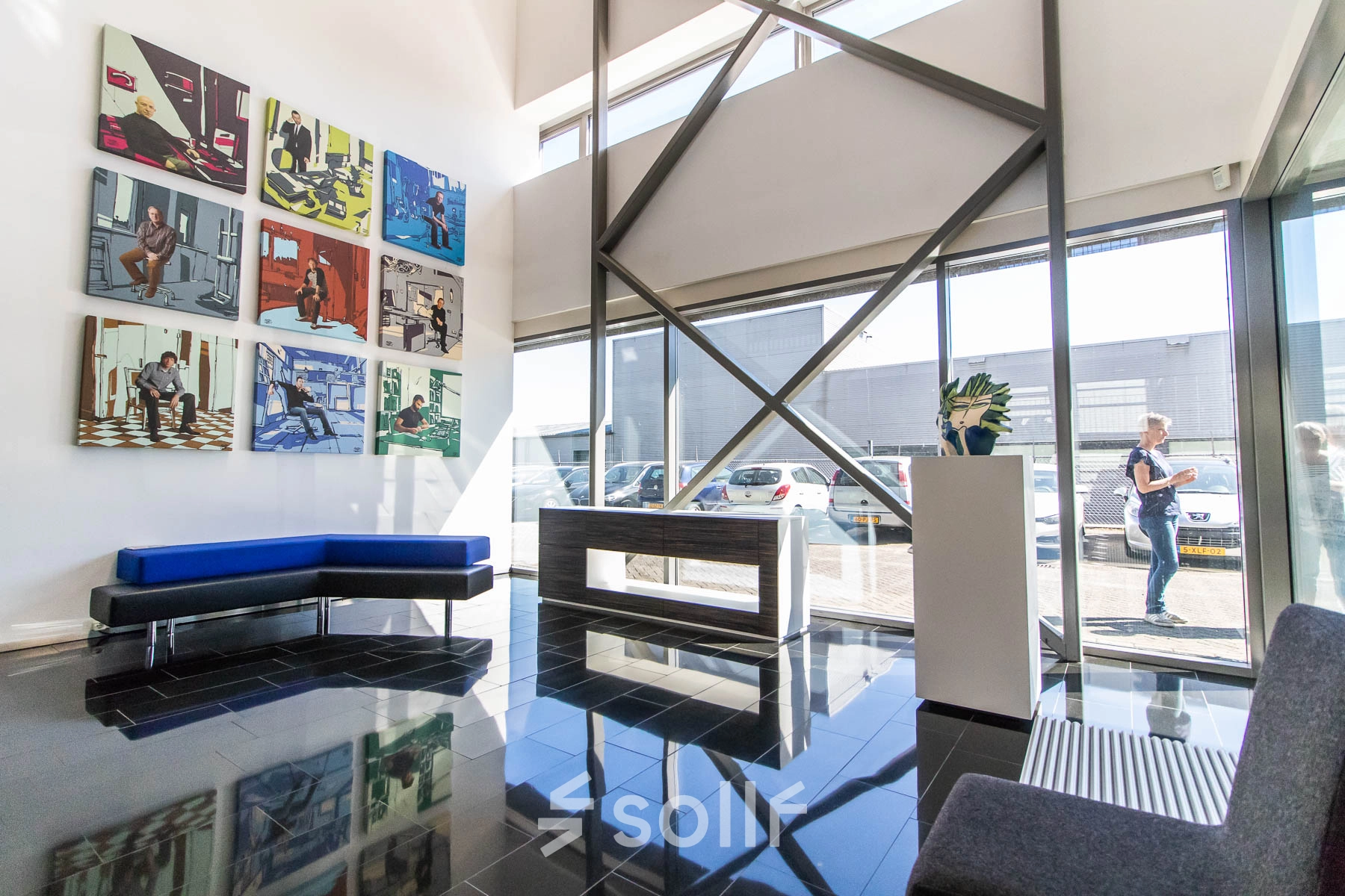 Bright office entrance with modern art on the wall, glass windows, and seating area. A person is walking outside. Ideal for office space rental.