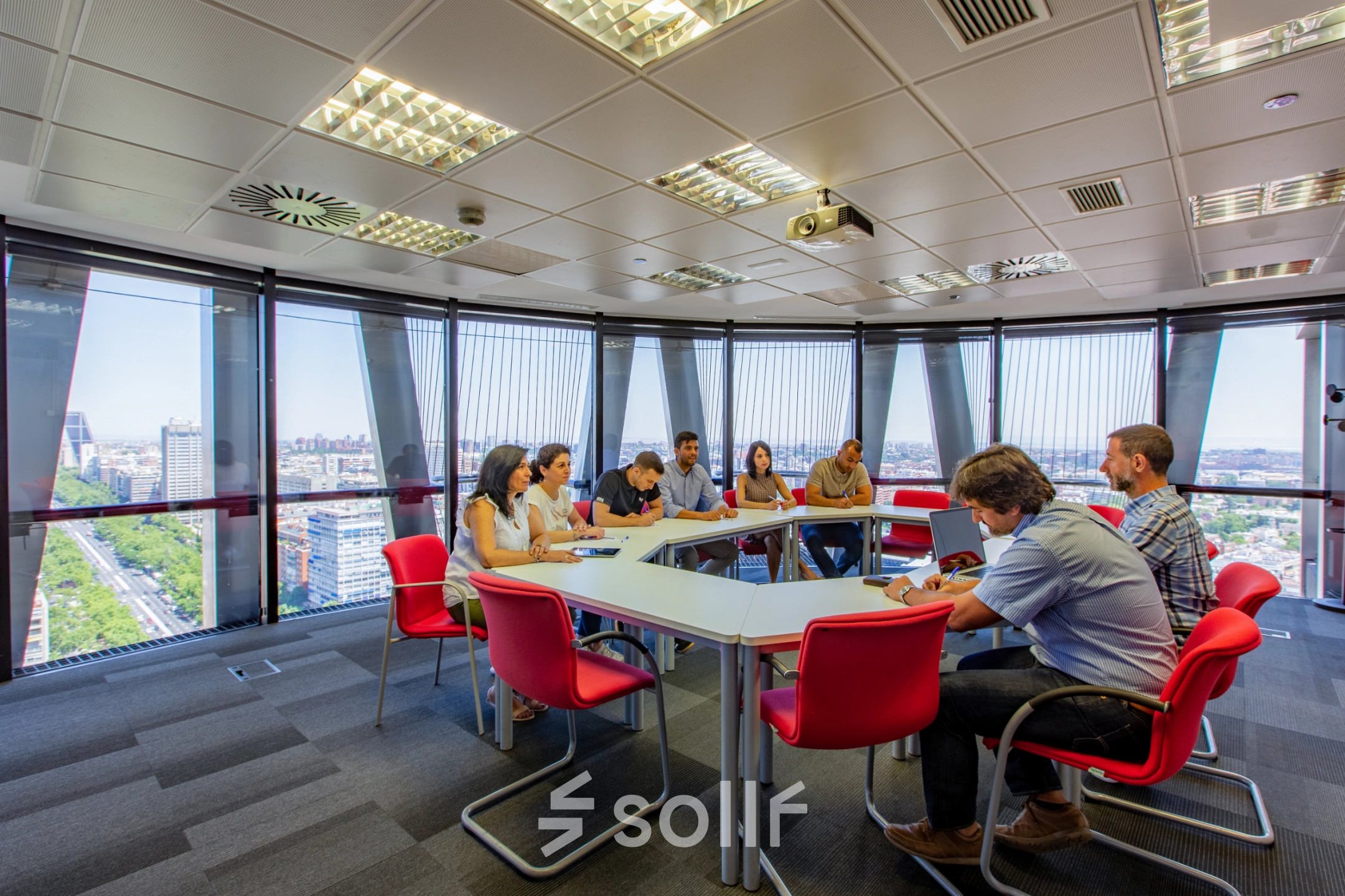 A conference room in Paseo de la Castellana 95, Madrid Azca, with people engaged in a meeting around a large table, featuring large windows and red chairs, available for office space rental.