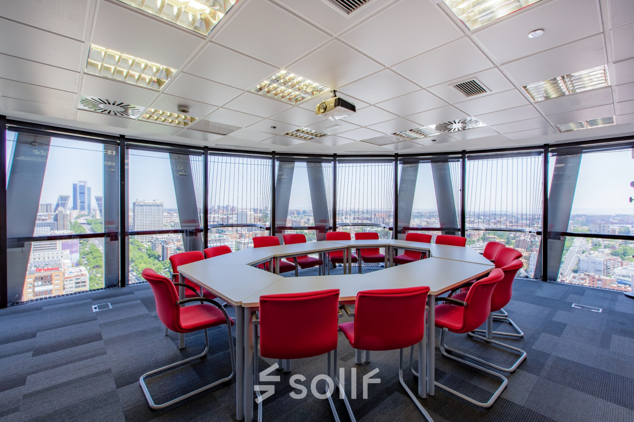 Bright office space with a panoramic city view, featuring a round conference table surrounded by red chairs, ideal for meetings or presentations.
