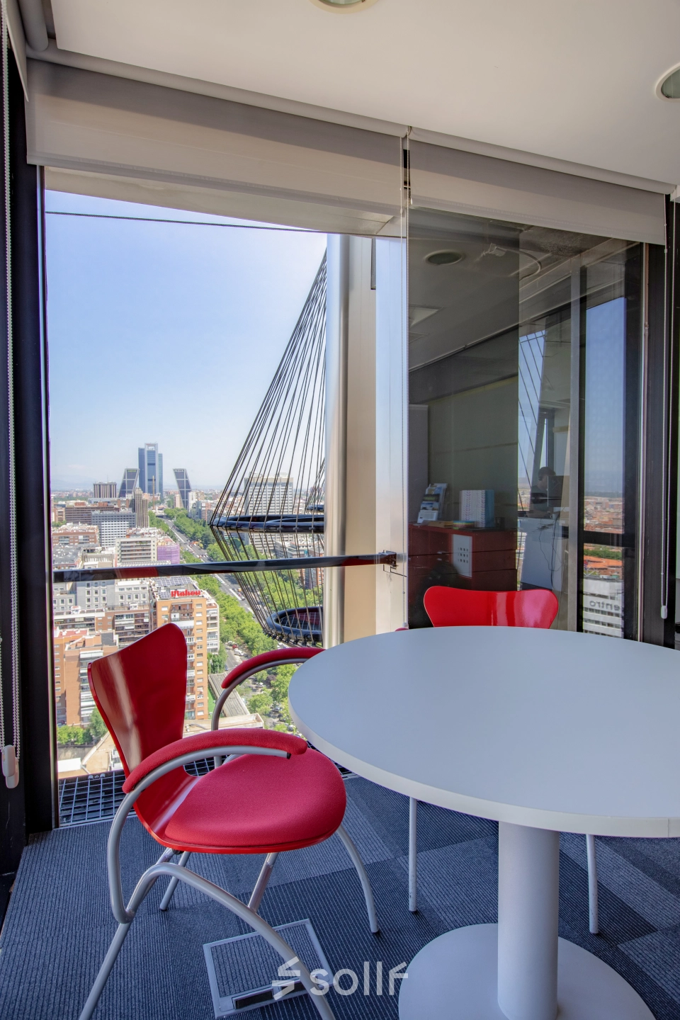 Office space for rent at Paseo de la Castellana 95, Madrid Azca, featuring a round white table and red chairs with a panoramic city view.