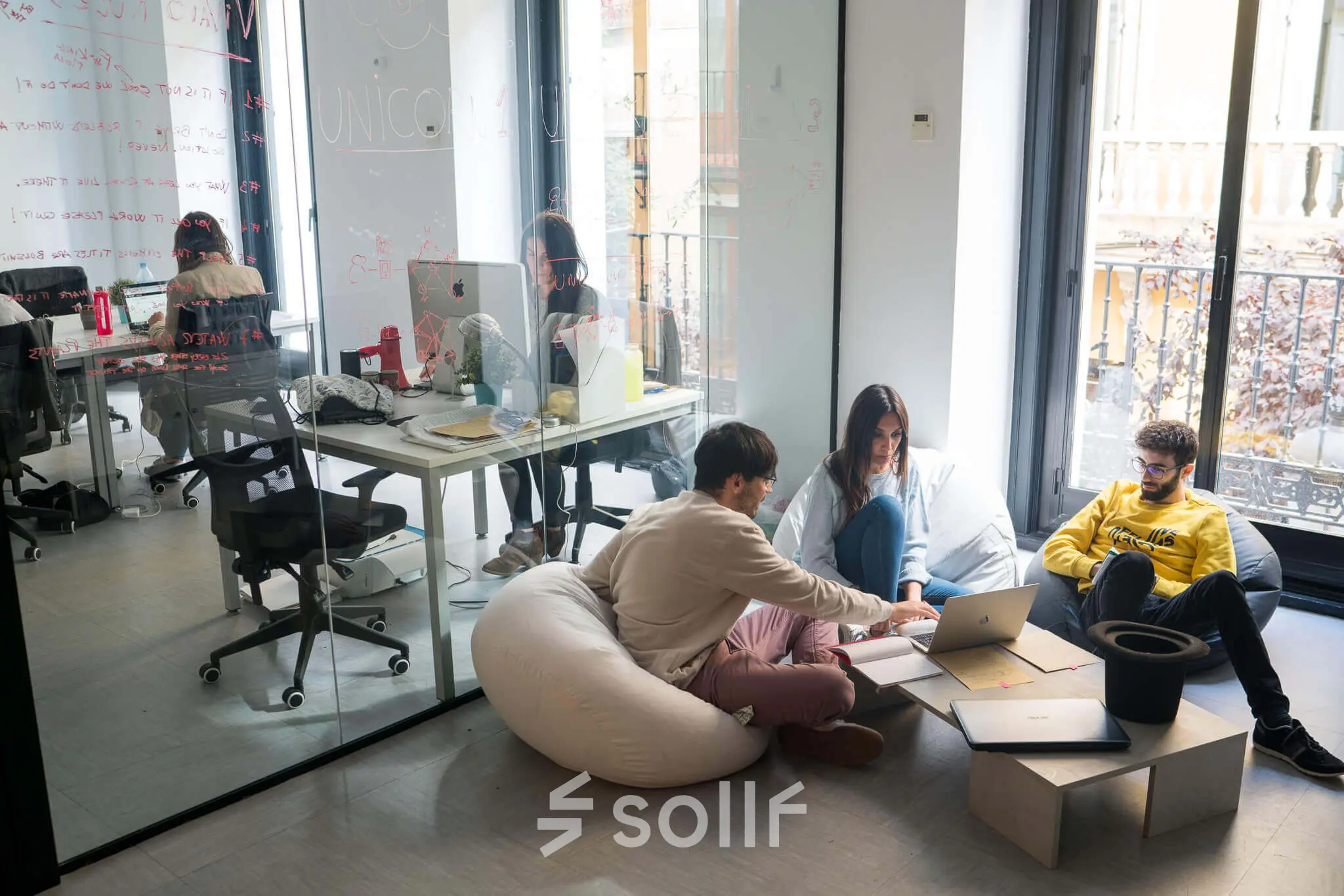 Office space to rent in Madrid Center with modern furnishings, featuring people collaborating around a laptop and others working at desks in a bright setting.