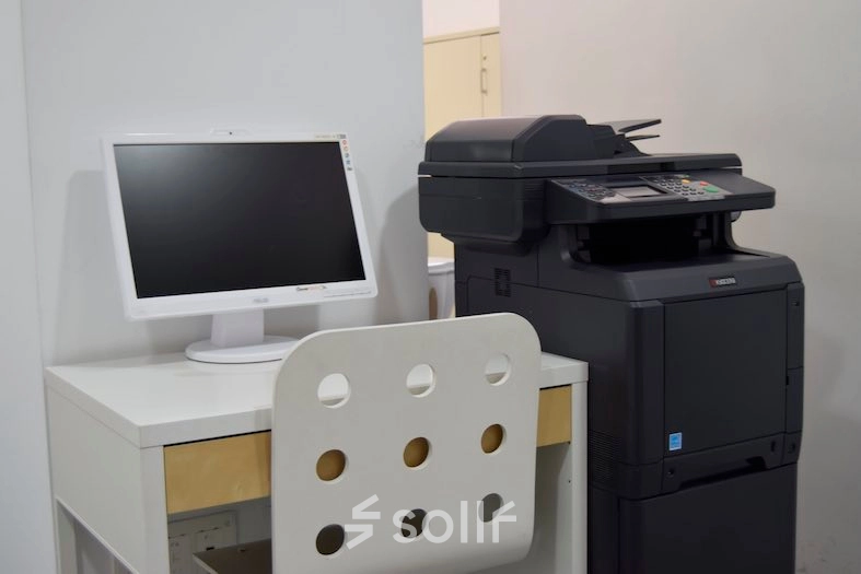 Compact office equipment setup with a desk, chair, monitor, and multifunction printer, ideal for those seeking office space rental in Madrid Center.