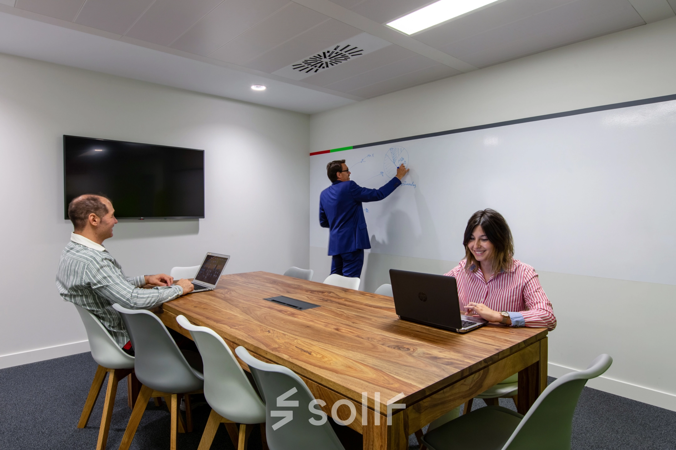 Professionals engage in business activities in a furnished office space with a conference table and a wall-mounted screen, ideal for office space rental in calle de O'Donnell, Madrid Retiro.