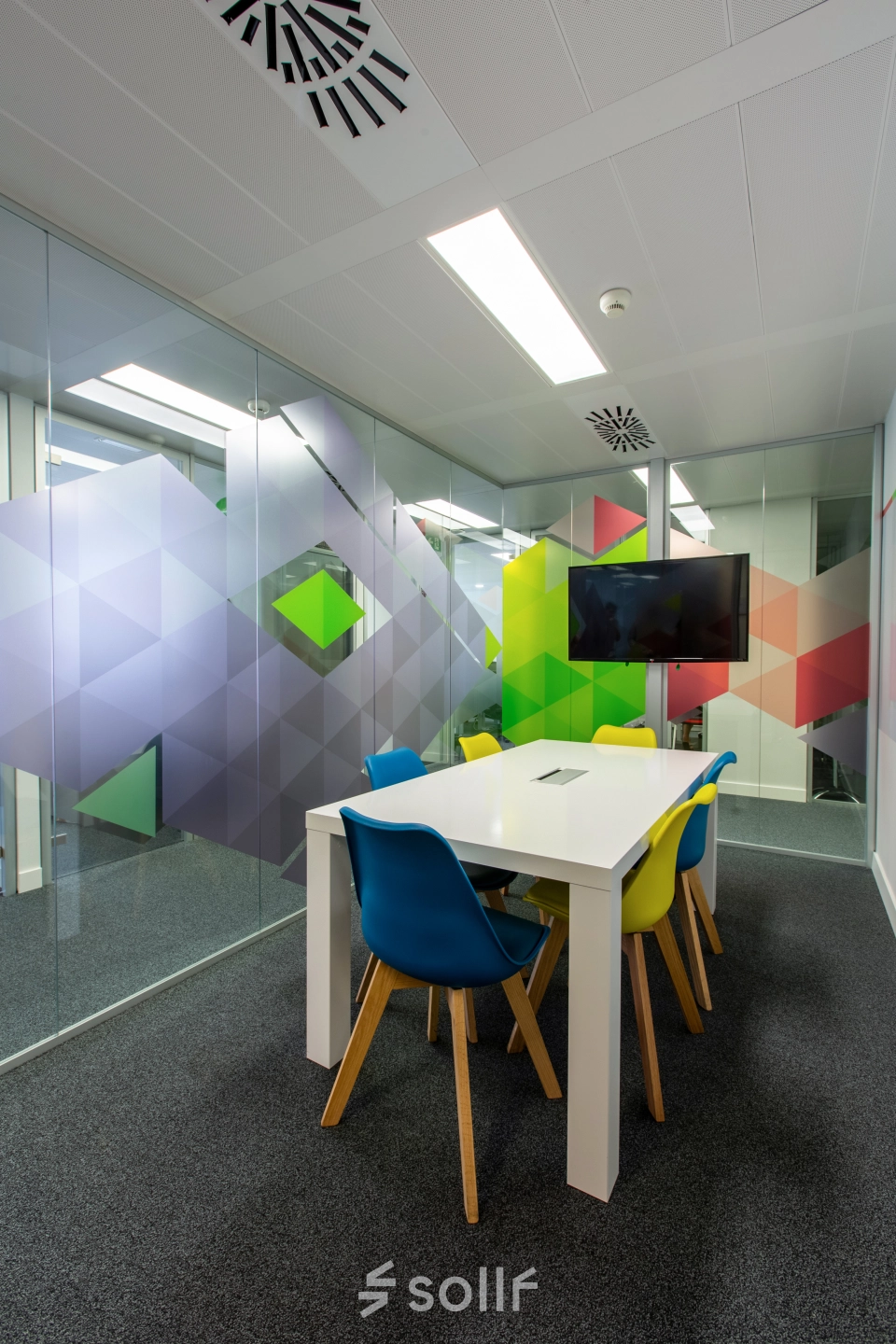 Modern office space rental at Calle de O'Donnell, Madrid Retiro. Features colorful chairs surrounding a white table in a meeting room with vibrant glass wall designs.