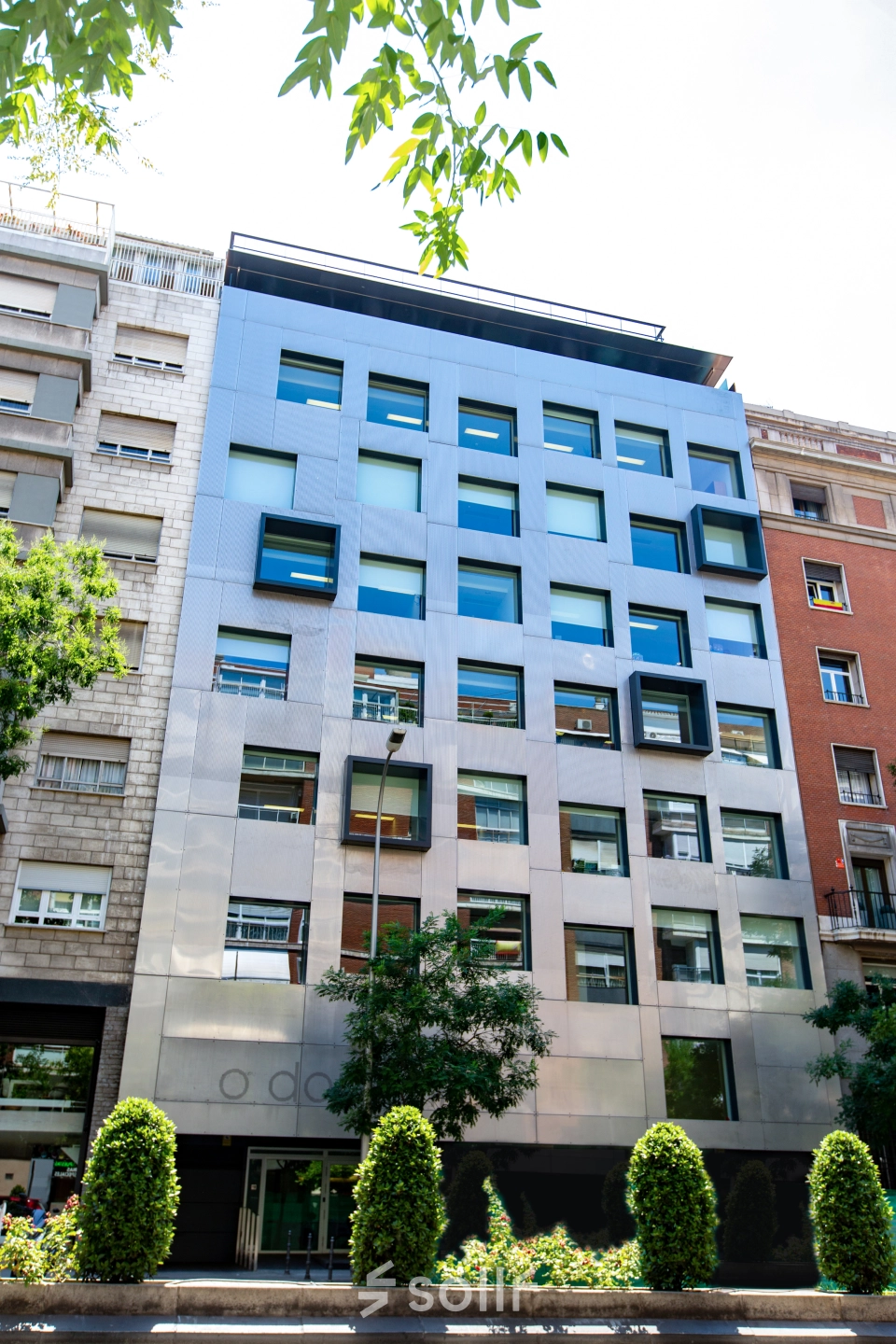 Modern exterior of an office building available for rent on calle de O'Donnell, Madrid Retiro, featuring geometric windows and surrounded by greenery.