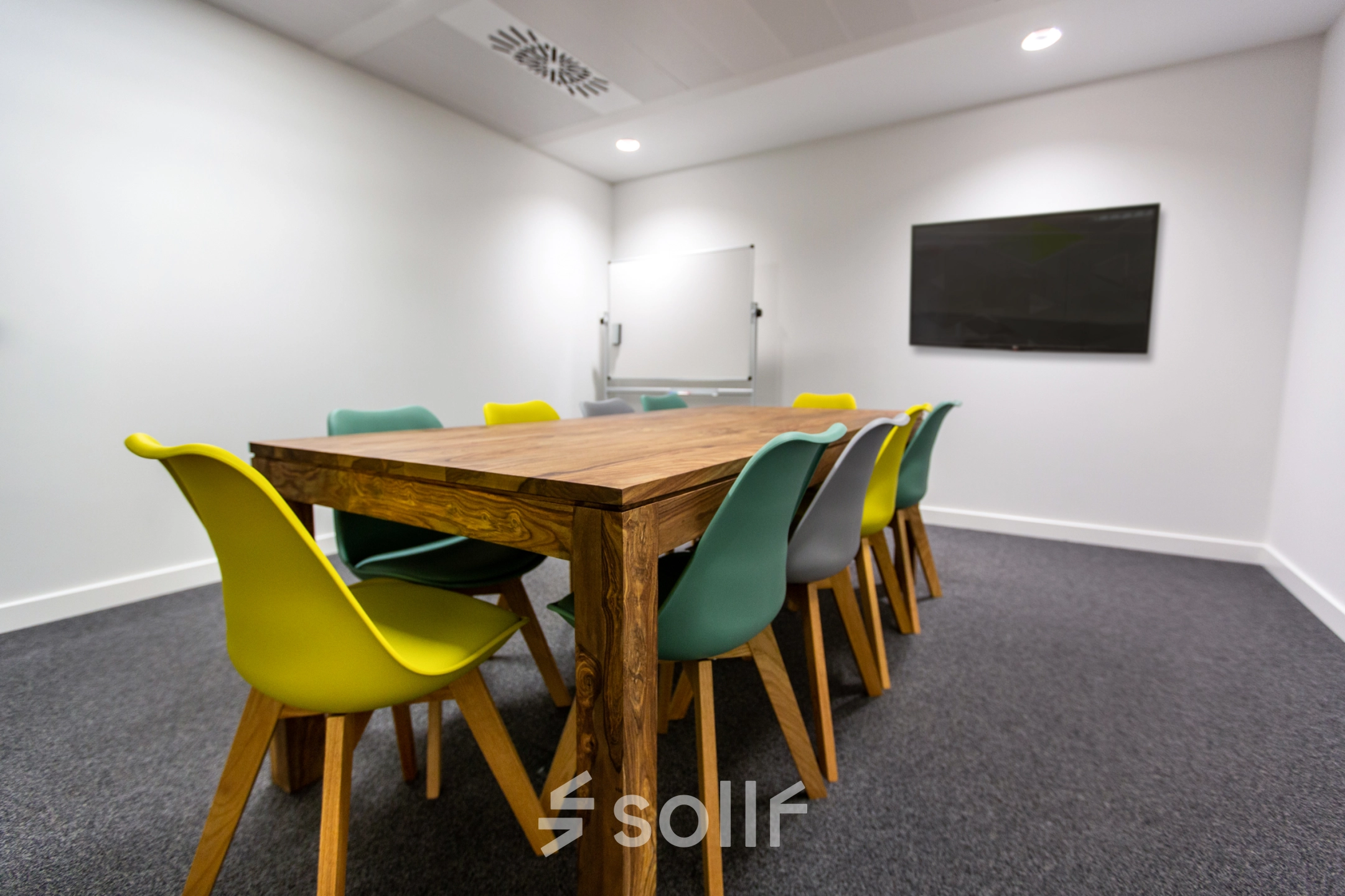 Modern office to rent in Madrid Retiro, featuring a wooden table with colorful chairs, whiteboard, and a screen, ideal for conferences or meetings.