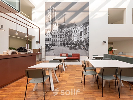 Bright office space in Mechelen Centrum with modern furniture and a large mural, ideal for office space rental. Tables and chairs arranged for collaborative work.