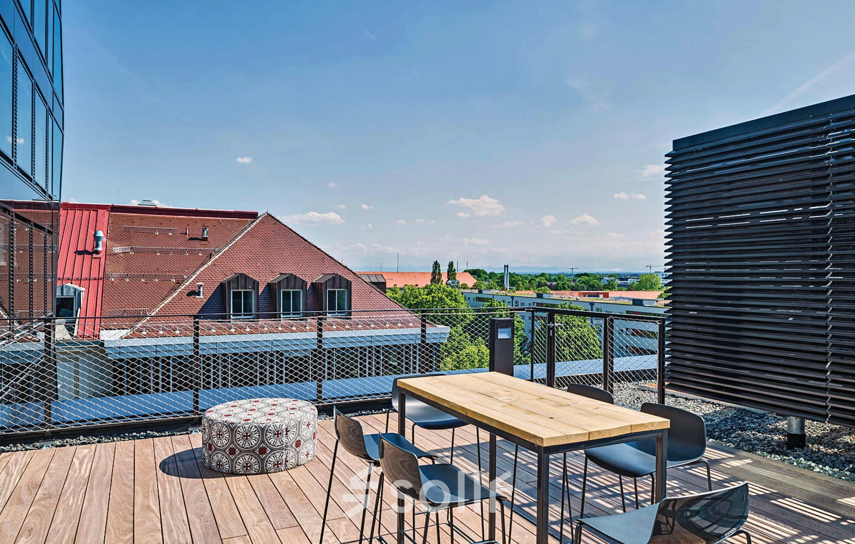 Outdoor office space in München Werksviertel with a wooden deck, table, and chairs offering a panoramic view of the surrounding rooftops and distant horizon.