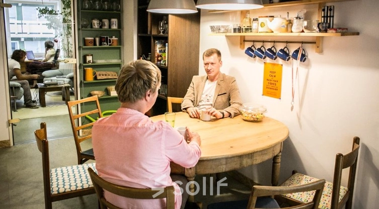 Two people are seated at a wooden table in a cozy, furnished office space, engaging in a conversation. The office includes shelves with cups and decorative items, available for office space rental.