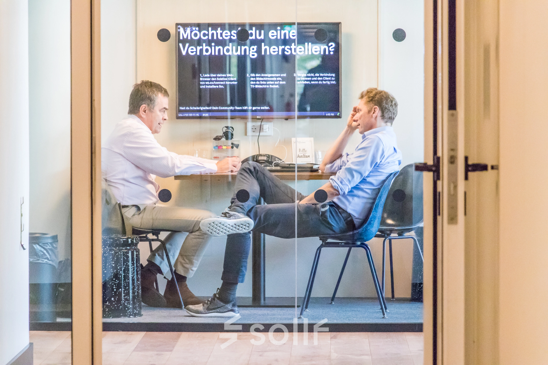Two people engaged in a discussion in a furnished office space with a digital screen on the wall in Oskar-von-Miller-Ring 20, Munich Maxvorstadt. Ideal for office space rental.