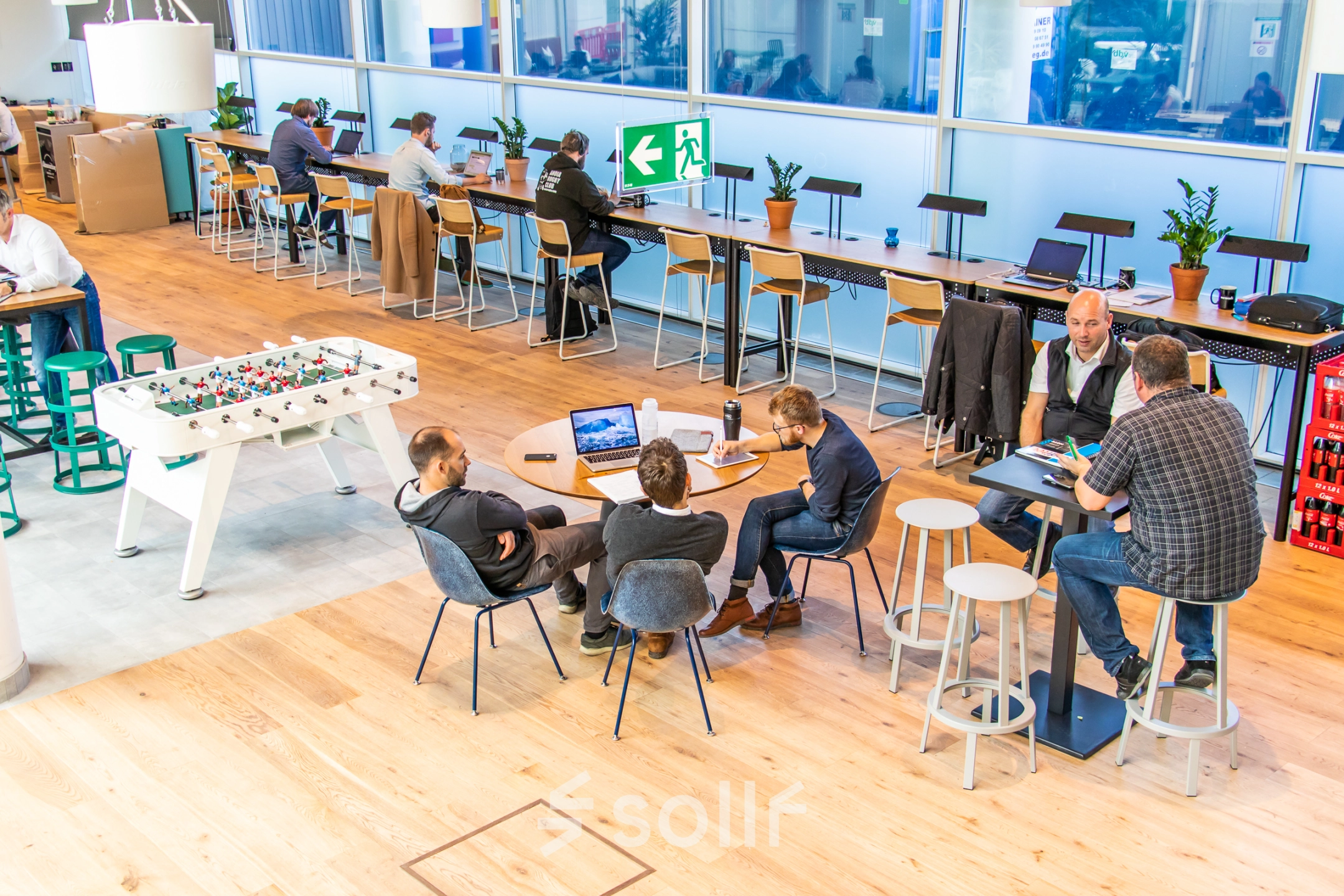 Bright, furnished workspace with people collaborating at tables, featuring long desks and a foosball table. Ideal for rent office space in Munich's Maxvorstadt neighborhood.