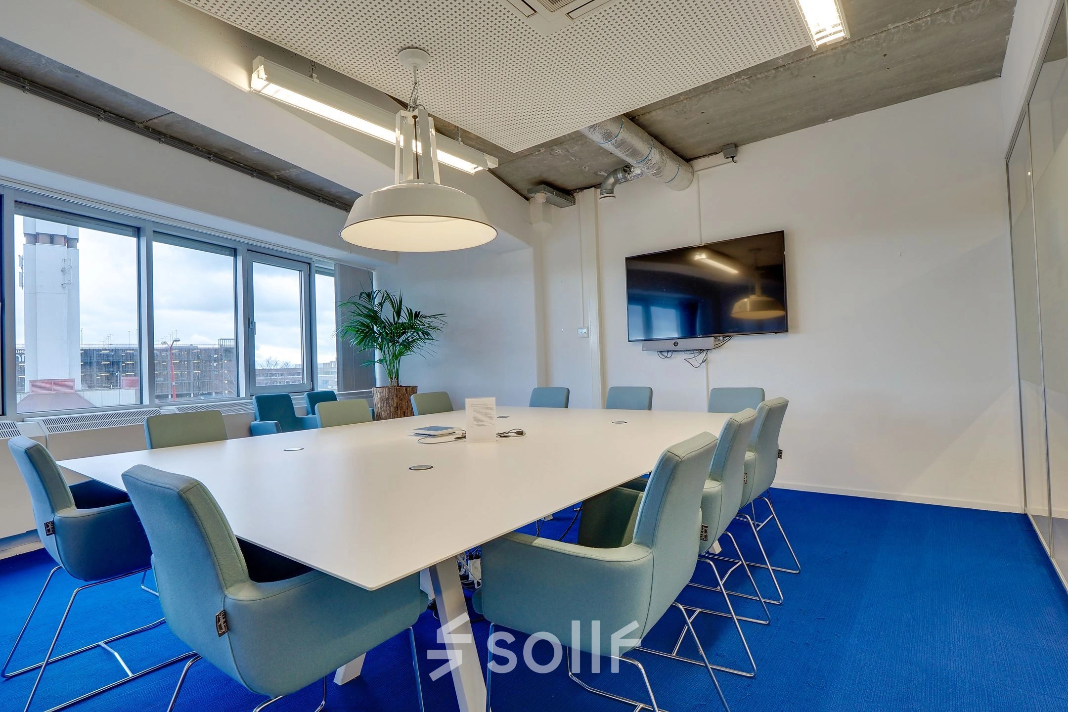 Modern conference room with a large white table surrounded by blue chairs, wall-mounted TV, and a window view at Zoomstede 19, Nieuwegein. Ideal for office space rental.