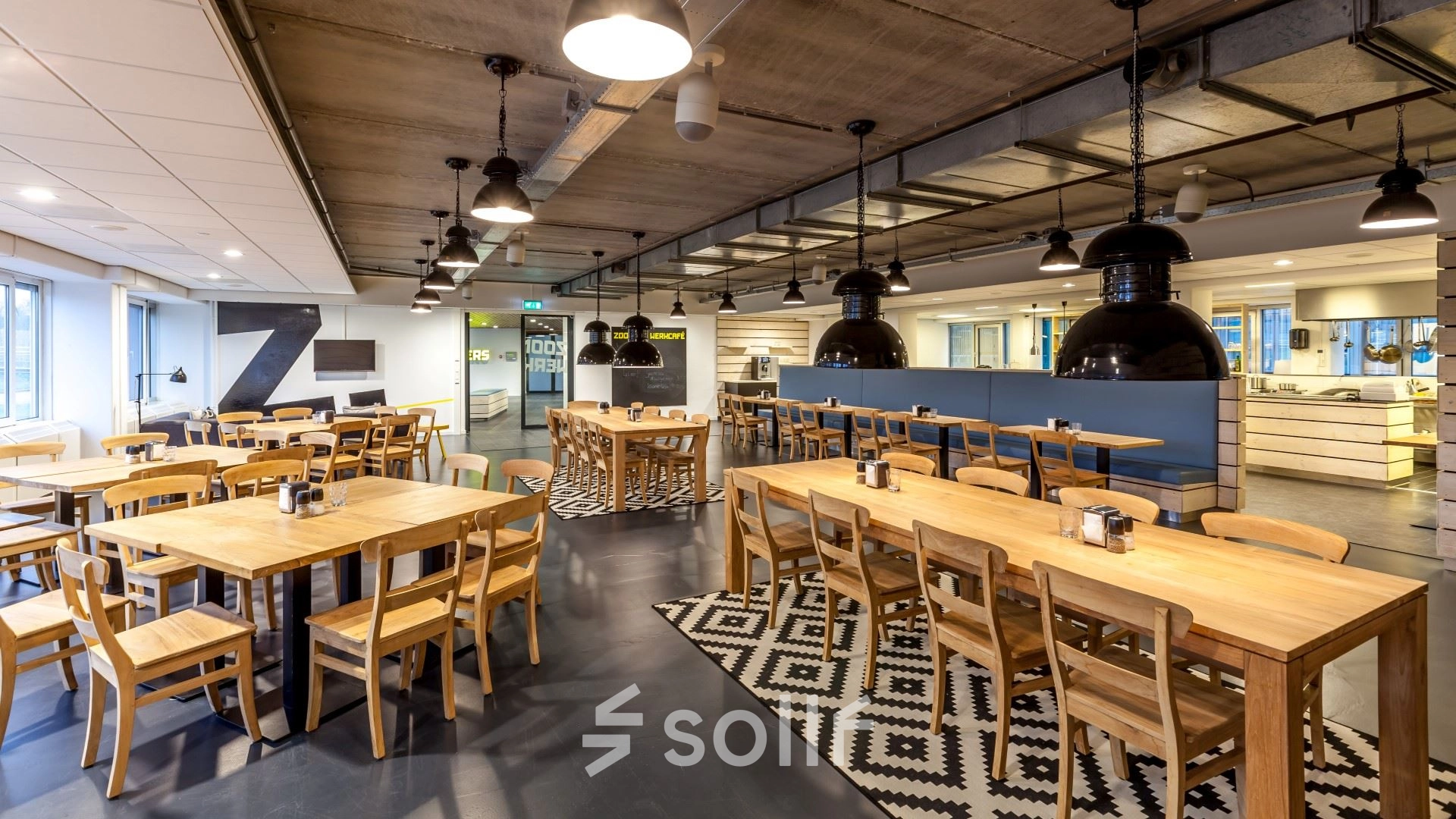 Spacious and modern interior with multiple wooden tables and chairs, perfect for collaborative work or dining. Bright overhead lighting and industrial-style decor add character to this inviting workspace. Ideal for office space rental.
