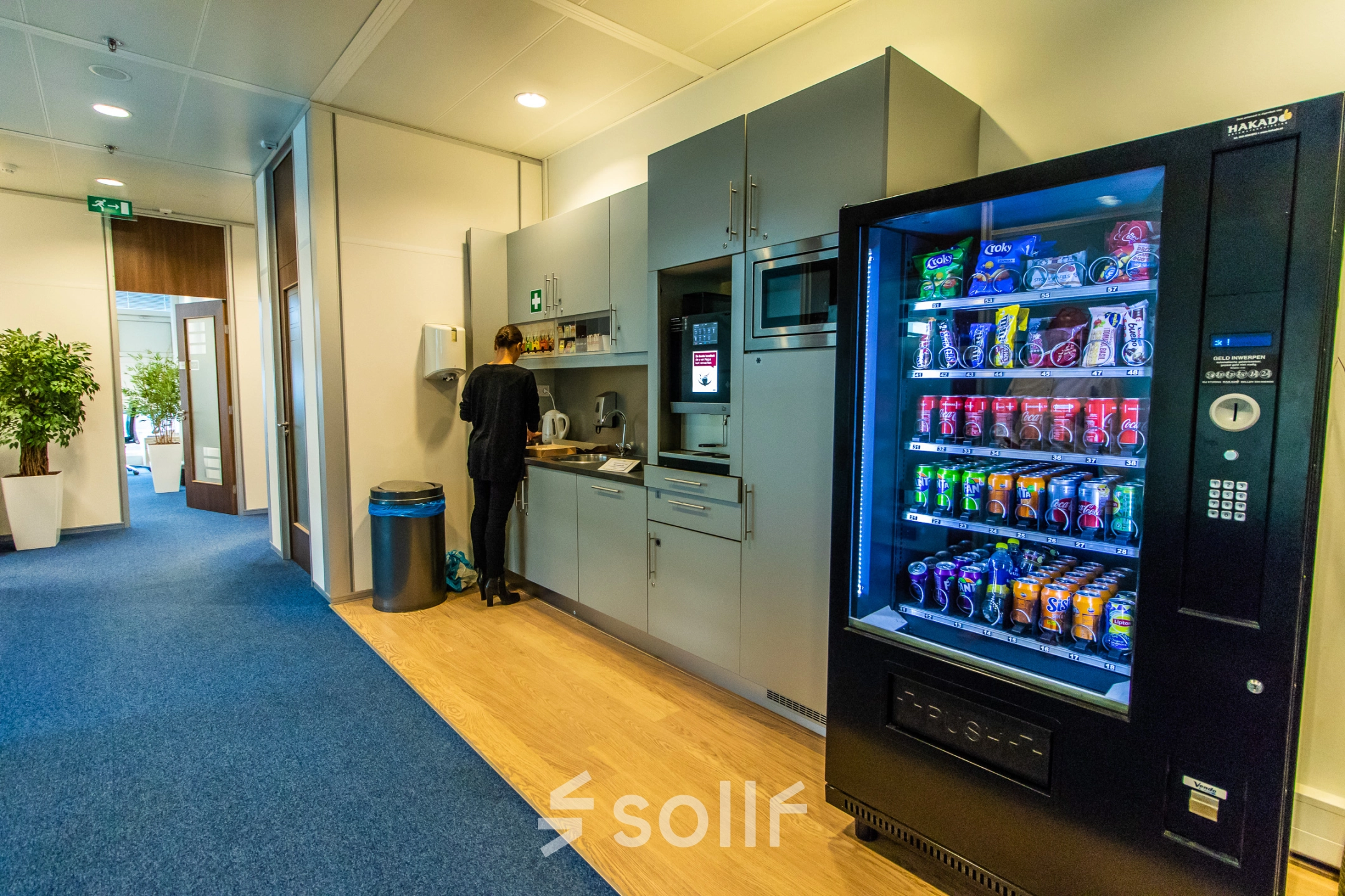 A furnished office space rental in Nijmegen featuring a pantry area with a person preparing a drink beside a stocked vending machine.