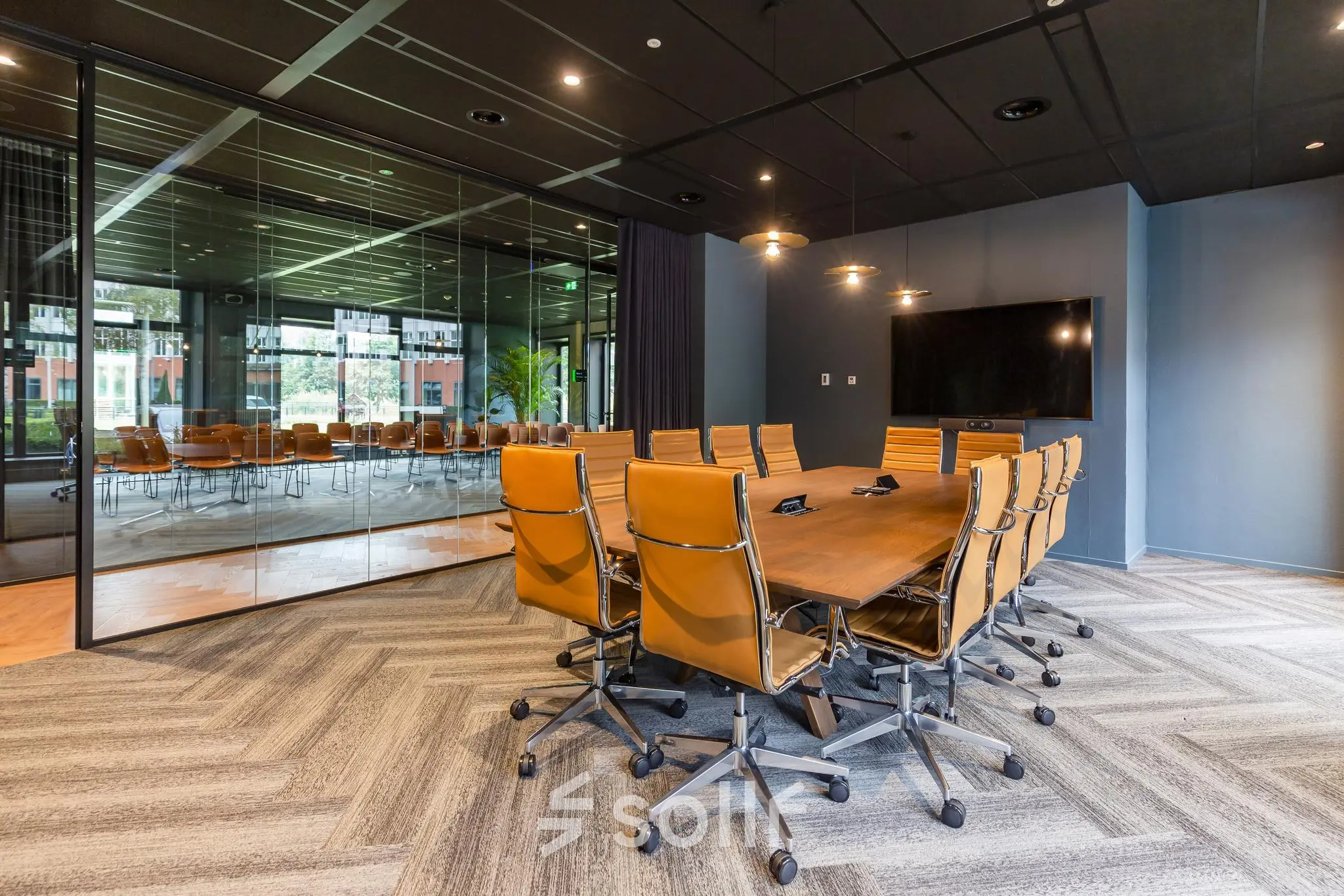 A modern conference room with a long wooden table and brown swivel chairs, equipped with video conferencing facilities. The glass wall reveals additional seating.