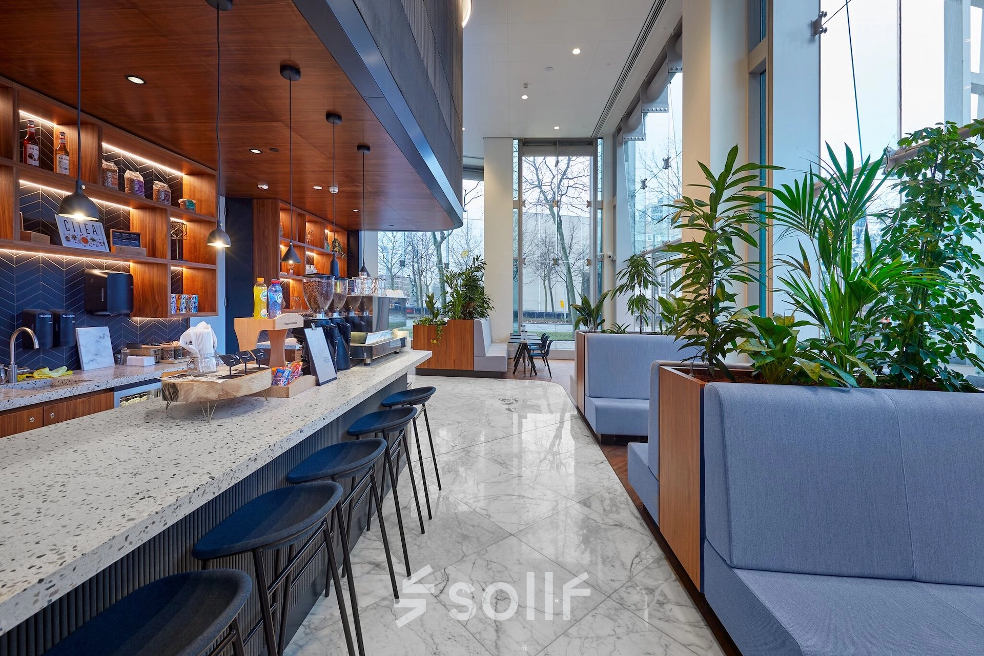 Modern office lounge area at Blaak 555 in Rotterdam Center with stylish seating, plants, and a coffee bar, ideal for office space rental.