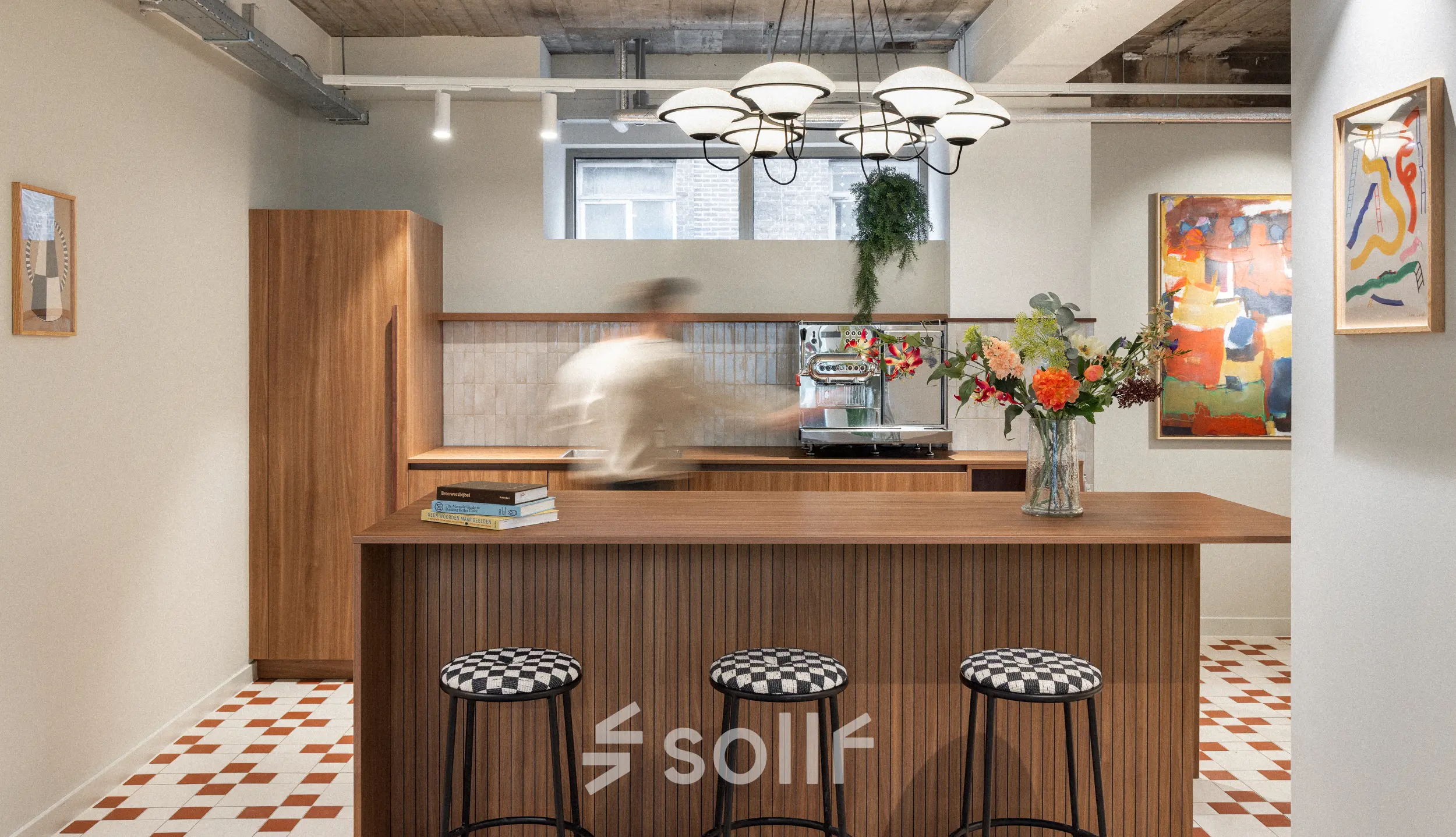 Stylish office lounge area with modern wooden furniture, art on the walls, and a person making coffee at the counter.