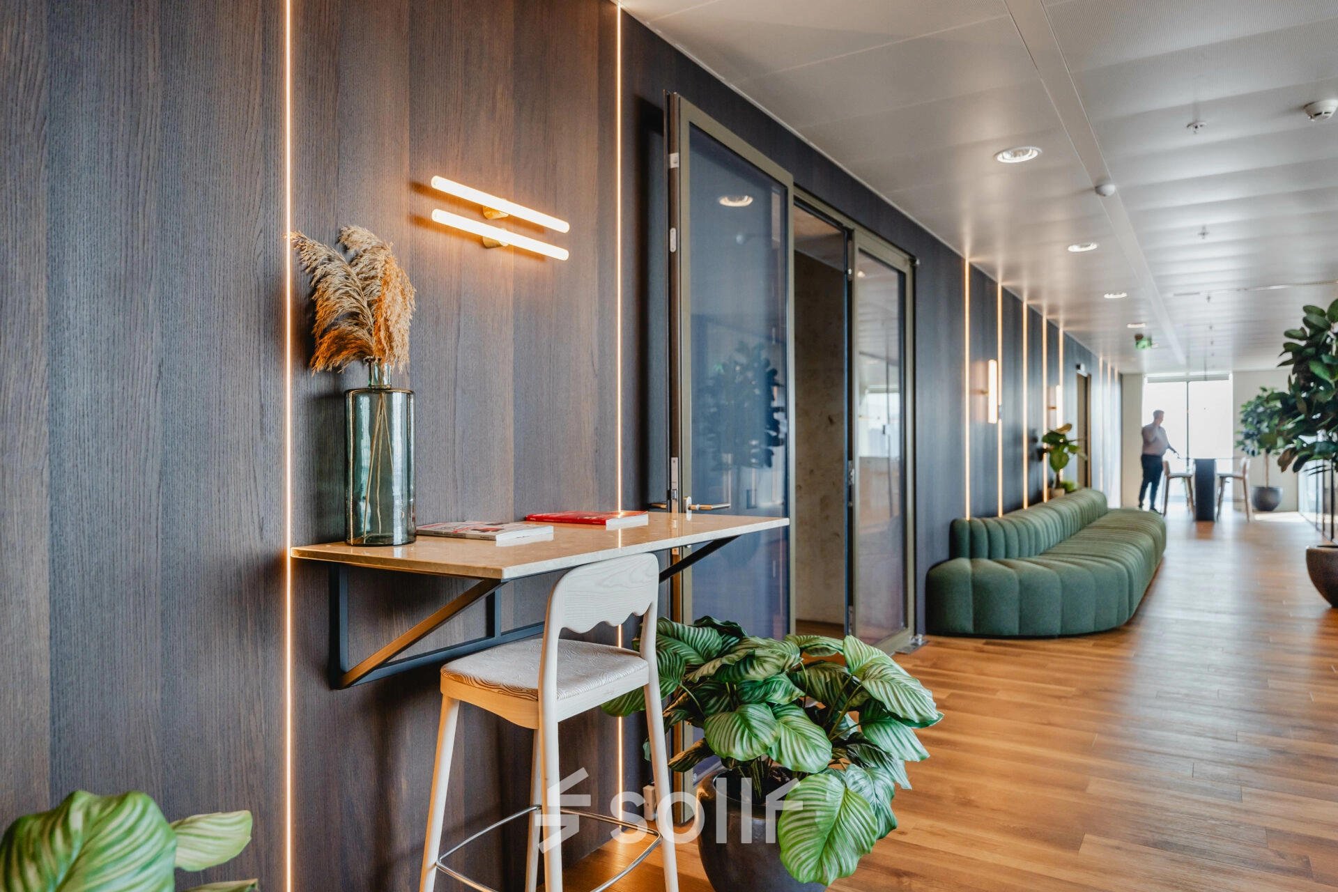 Stylish office at Weena 788 in Rotterdam Center featuring modern wooden accents, greenery, and a compact workspace ideal for rent. A person is seen engaging in business activities in the background.